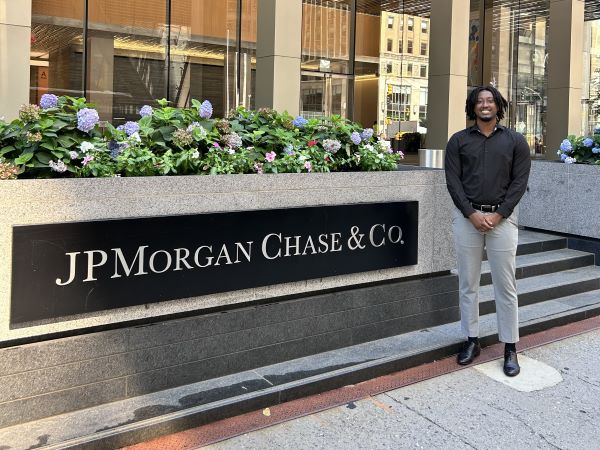 Lee Rwema in business casual stands next to glass building of JPMorganChase in NYC