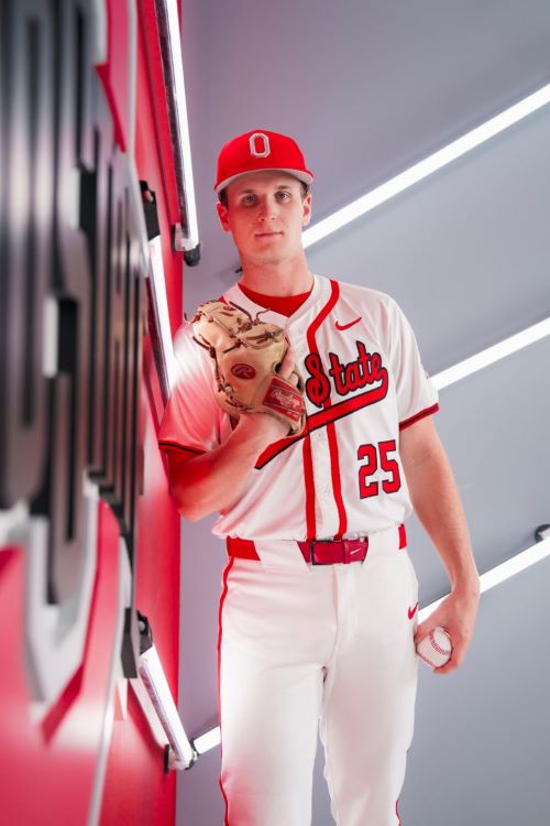 Portrait of Luke Carrell in his number 25 white with red lettering Ohio State baseball uniform, leaning against an Ohio State logo