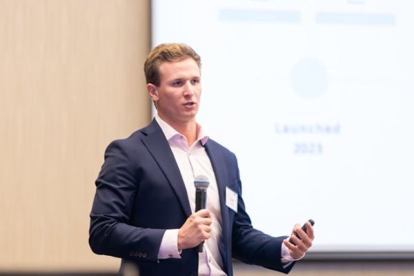 Luke Carrell in Blue suitcoat holding microphone with screen showing his presentation in the background