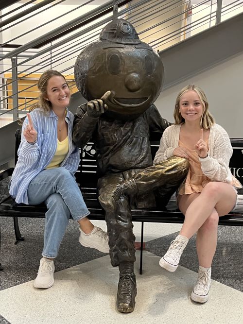 Two women sit on a bench with a bronze statue of Brutus Buckeye