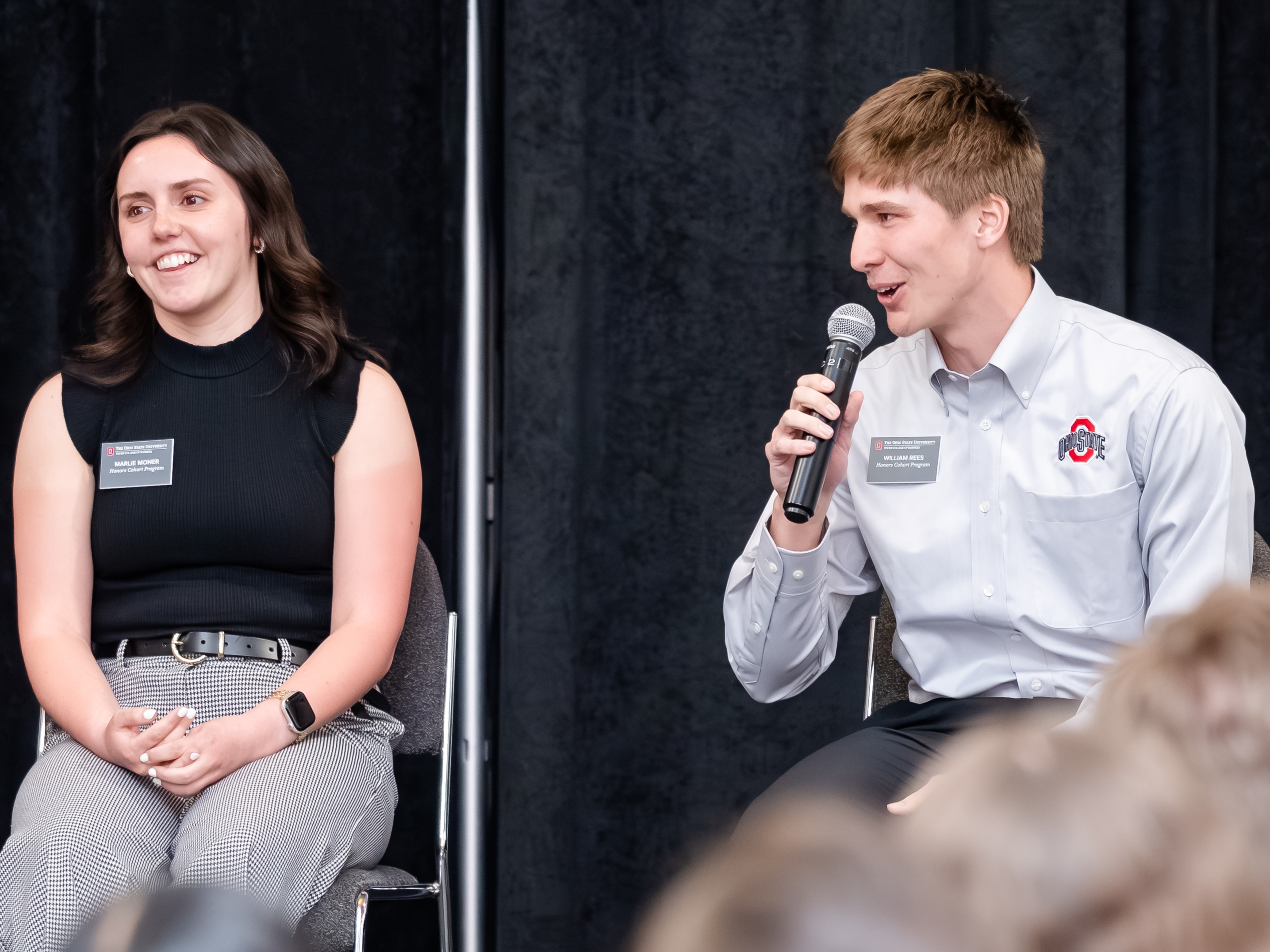 Students Marlie Moner, left, and Will Rees, holding a microphone, are seated on a stage with a black backdrop speaking to an audience of alumni.