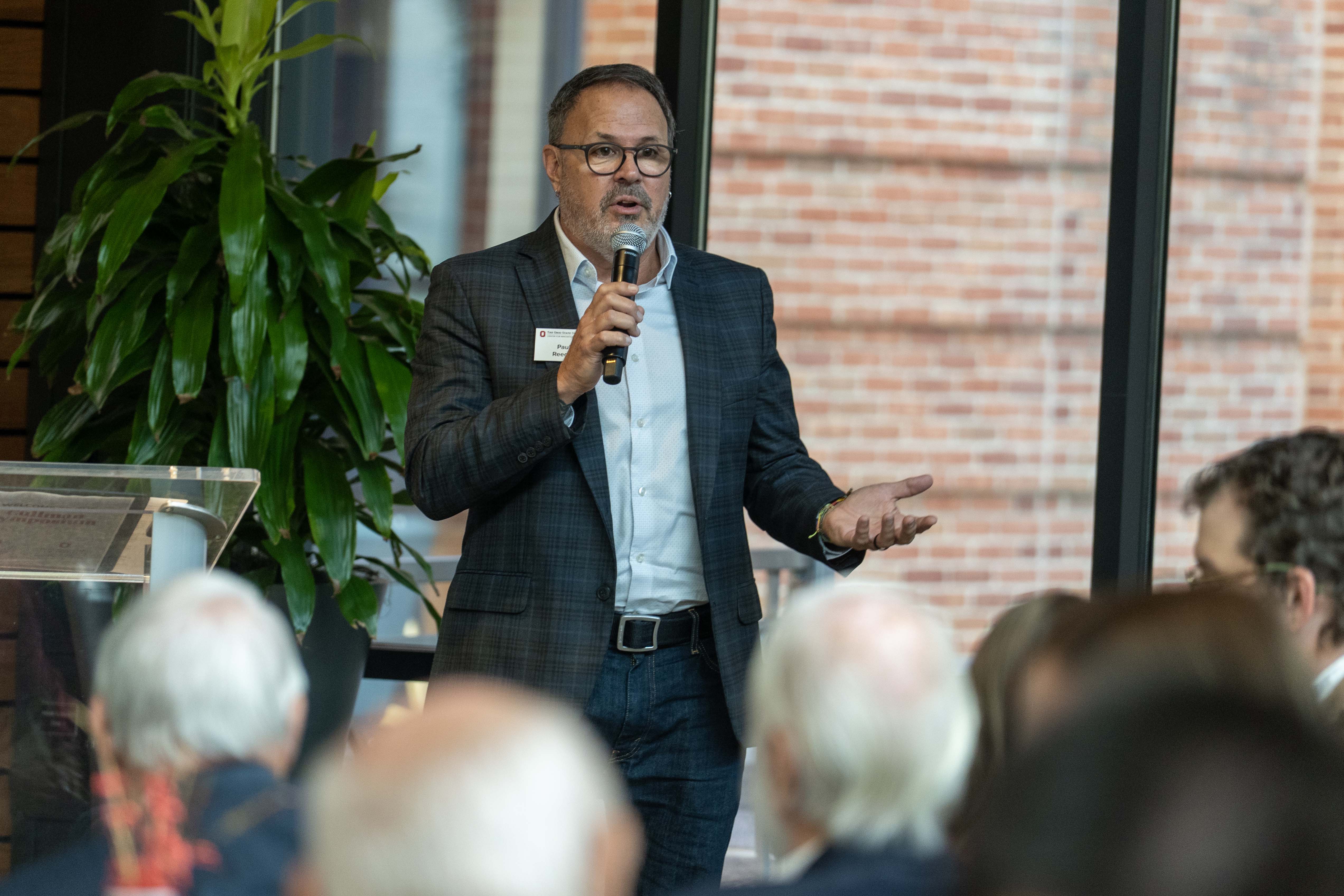 Paul Reeder in a suit jacket and jeans speaks into a microphone in front of a seated audience.