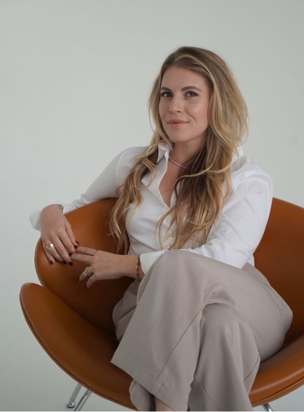 Sophia Mullins in tan slacks and a white blouse is seated in a modern brown leather chair.
