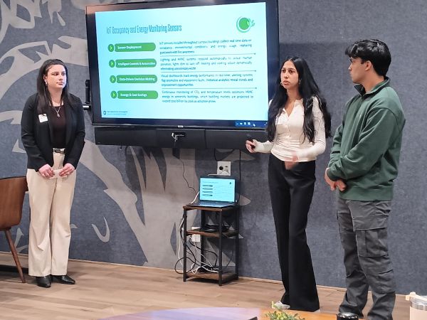 Three students in front of room describing what's on their PowerPoint projected on a screen
