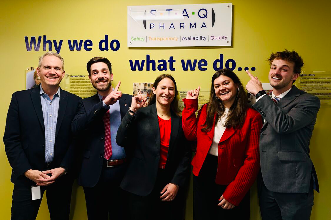 Students in business suits pose in front of a STAQ Pharma sign.