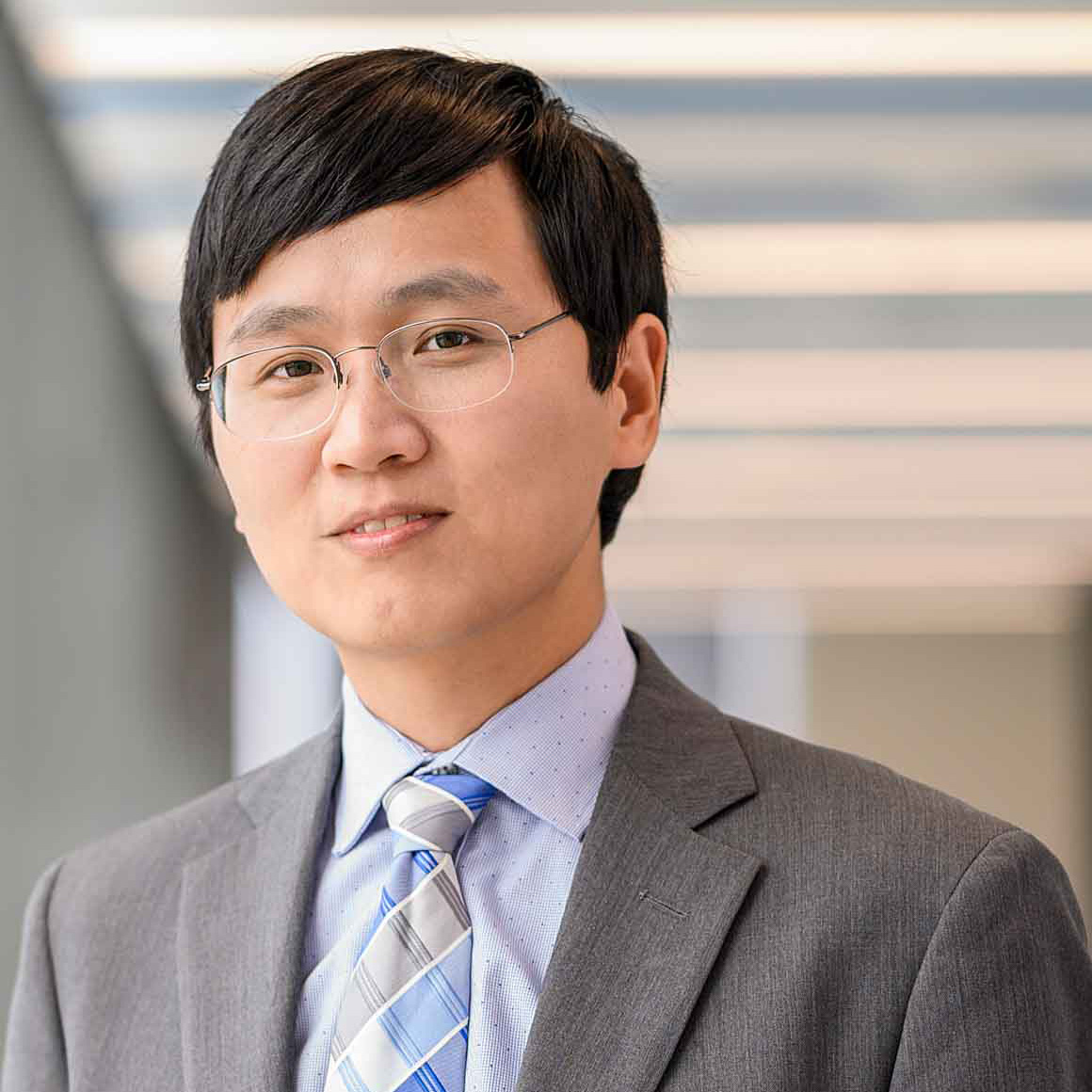Headshot of Tinglong Dai wearing glasses, a gray suit jacket, light blue shirt and blue patterned tie, standing indoors against a softly blurred background.
