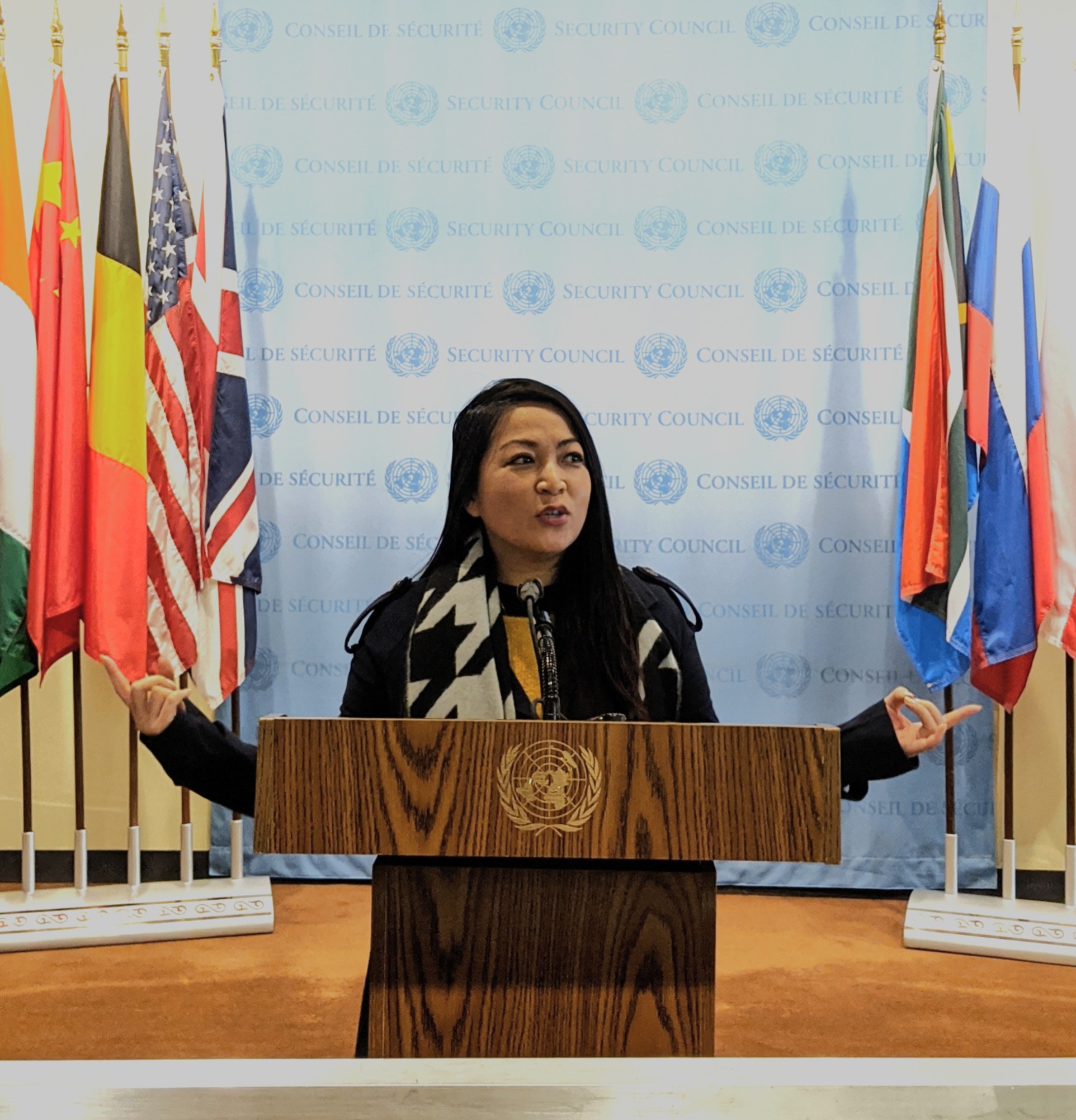 Sera standing behind a podium and speaking into a microphone with flags representing various nations behind her.