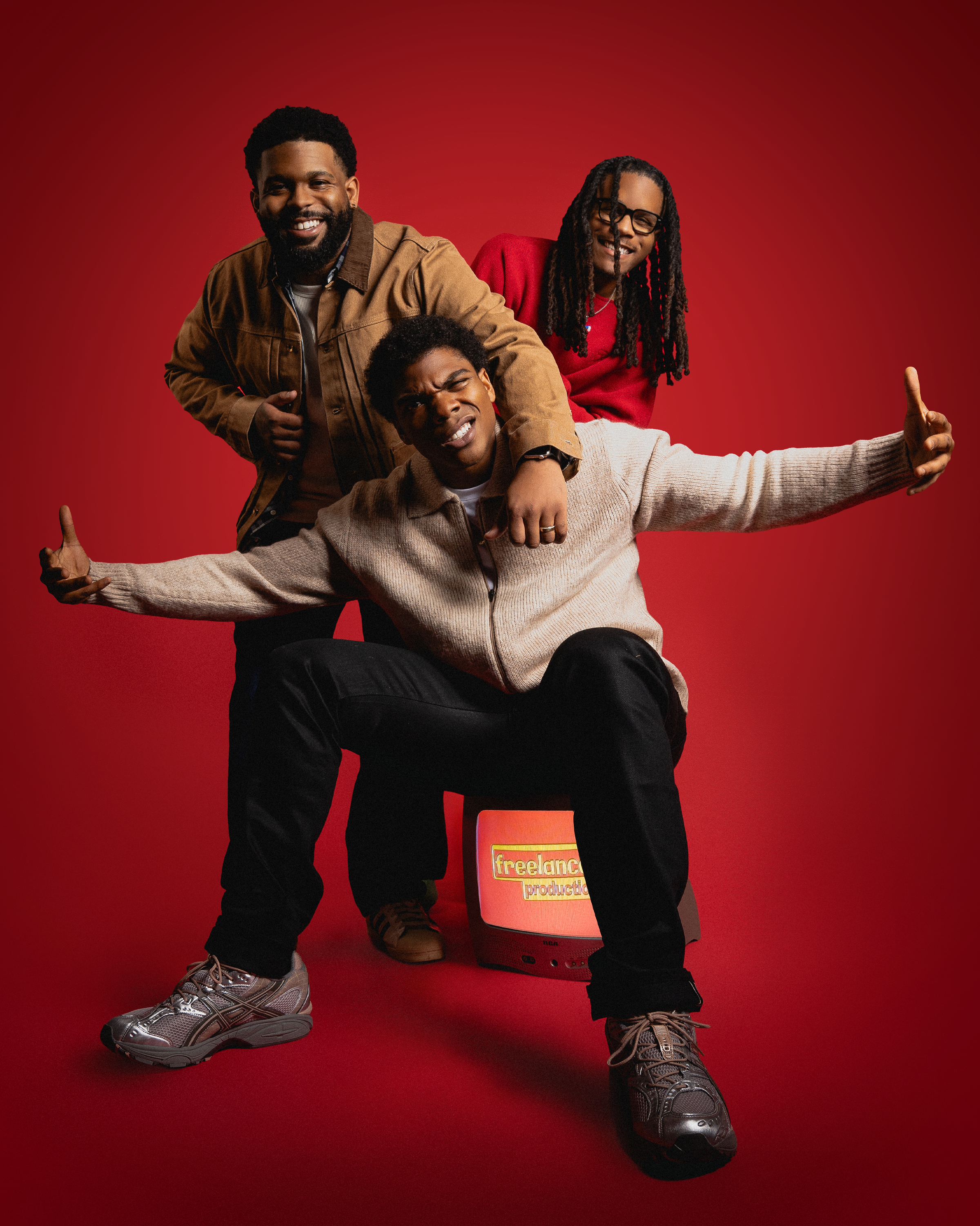 Against a red backdrop, Julien and Justen Turner stand behind actor Spence Moore who is seated and has his arms outstretched.