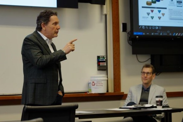 Lecturer stands in front of whiteboard in class while seated professor looks on
