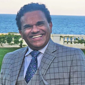 Headshot of Jojy Mathew in a gray suit with water in the background.