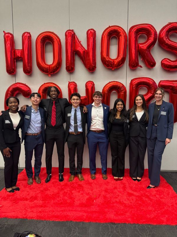 Group of eight students with arms on each other's shoulders stand on red carpet with the words Honors Cohort behind them