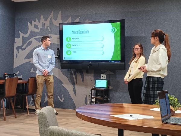 Three students in front of room explaining a PowerPoint on a pull-down screen