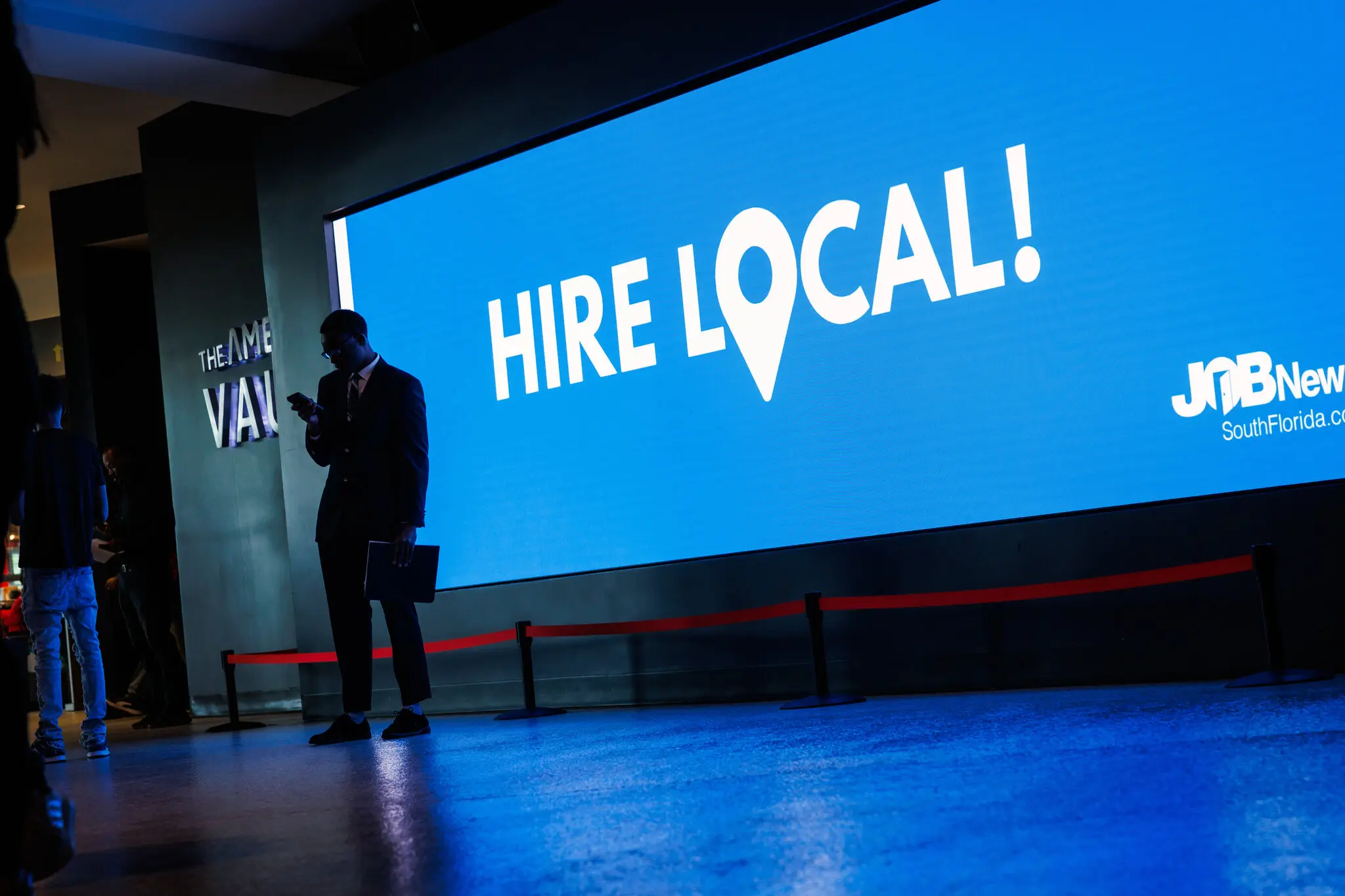 A man checks his phone in front of a blue monitor that says "Hire Local!"