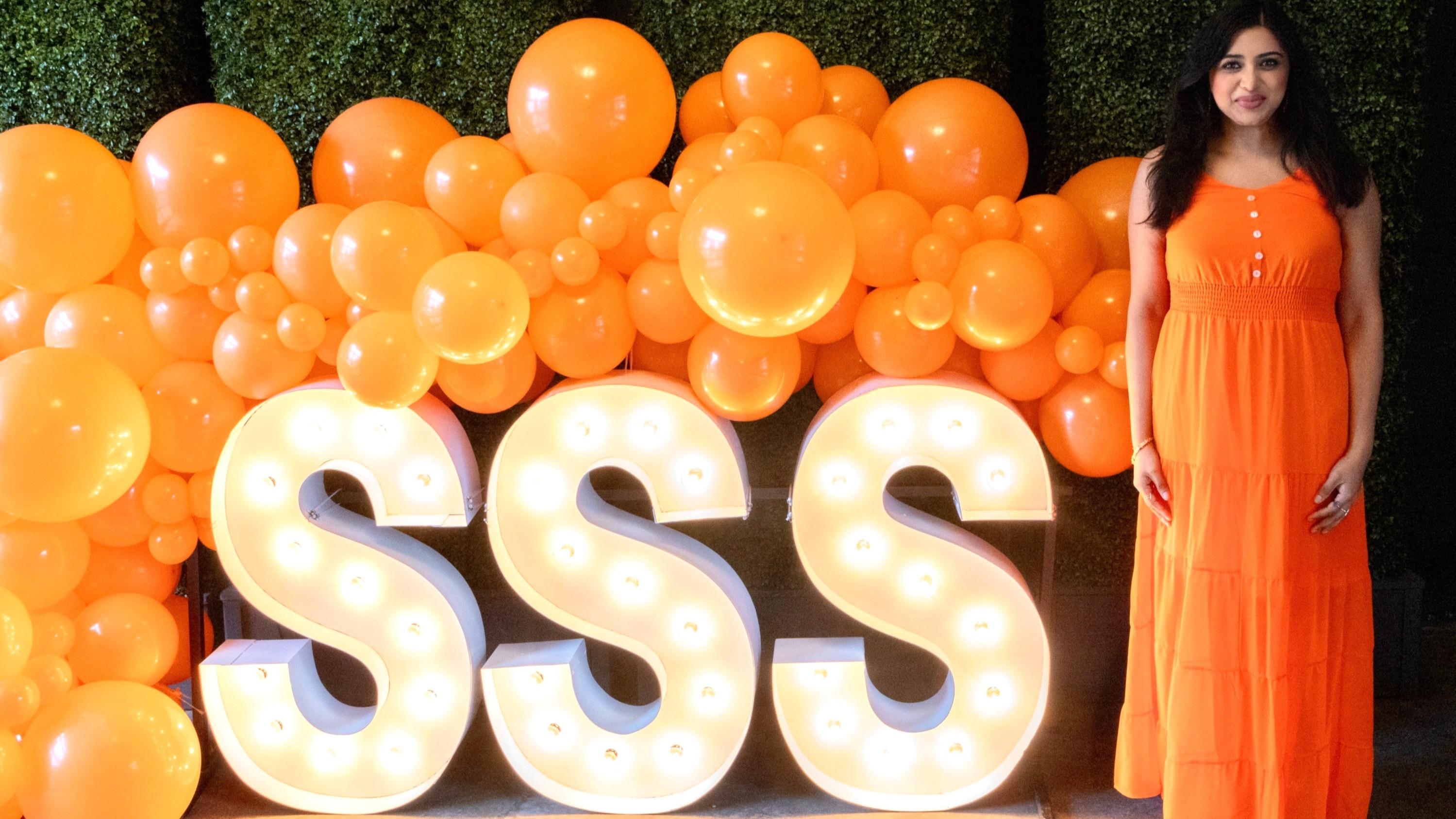Neethi Johnson in an orange dress standing to the right of a lighted SSS sign and decorative orange balloons.