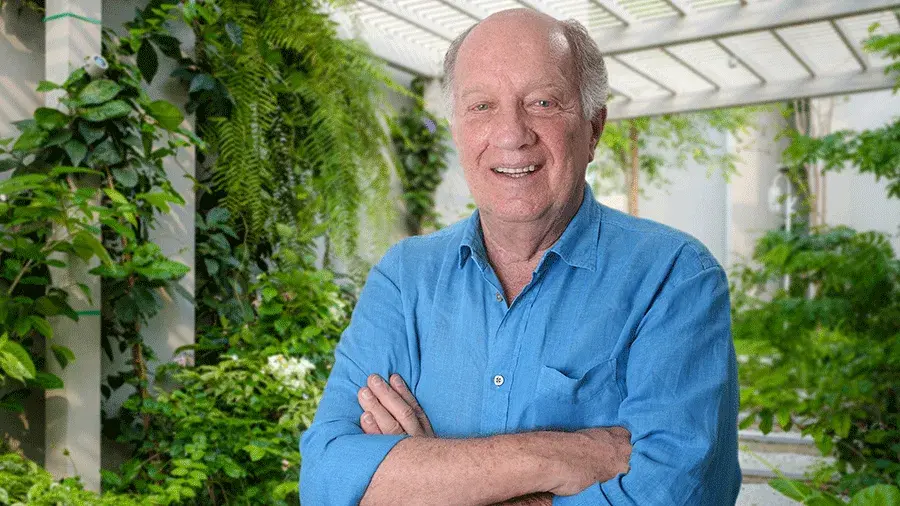 Roberto Abramson poses with arms folded in a blue shirt with green plants in the background.
