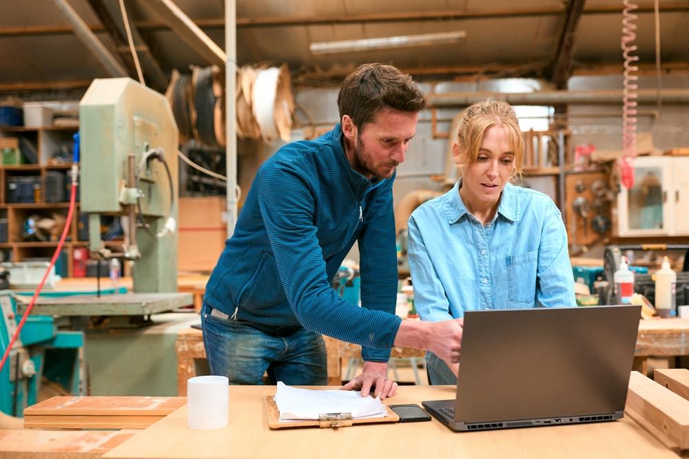 Two people in a workshop looking at a laptop on a wooden table, surrounded by woodworking tools and equipment.