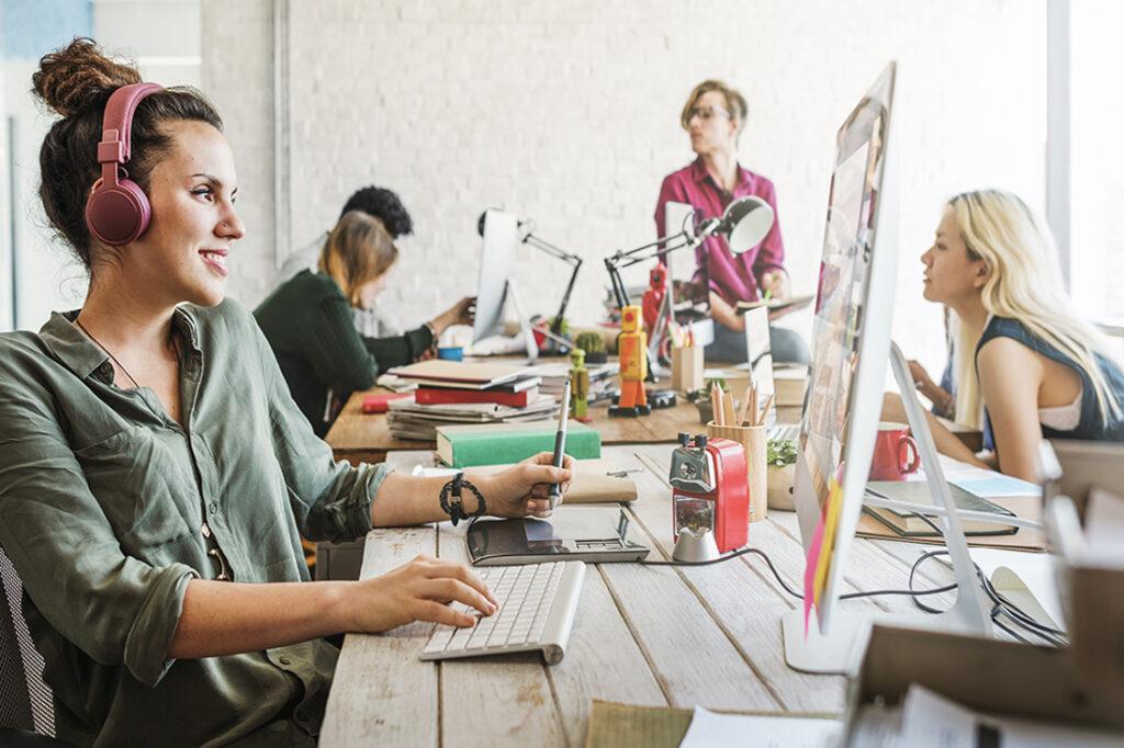 A smiling woman wearing pink headphones works on a computer in a bright, modern office with several coworkers collaborating and talking in the background.