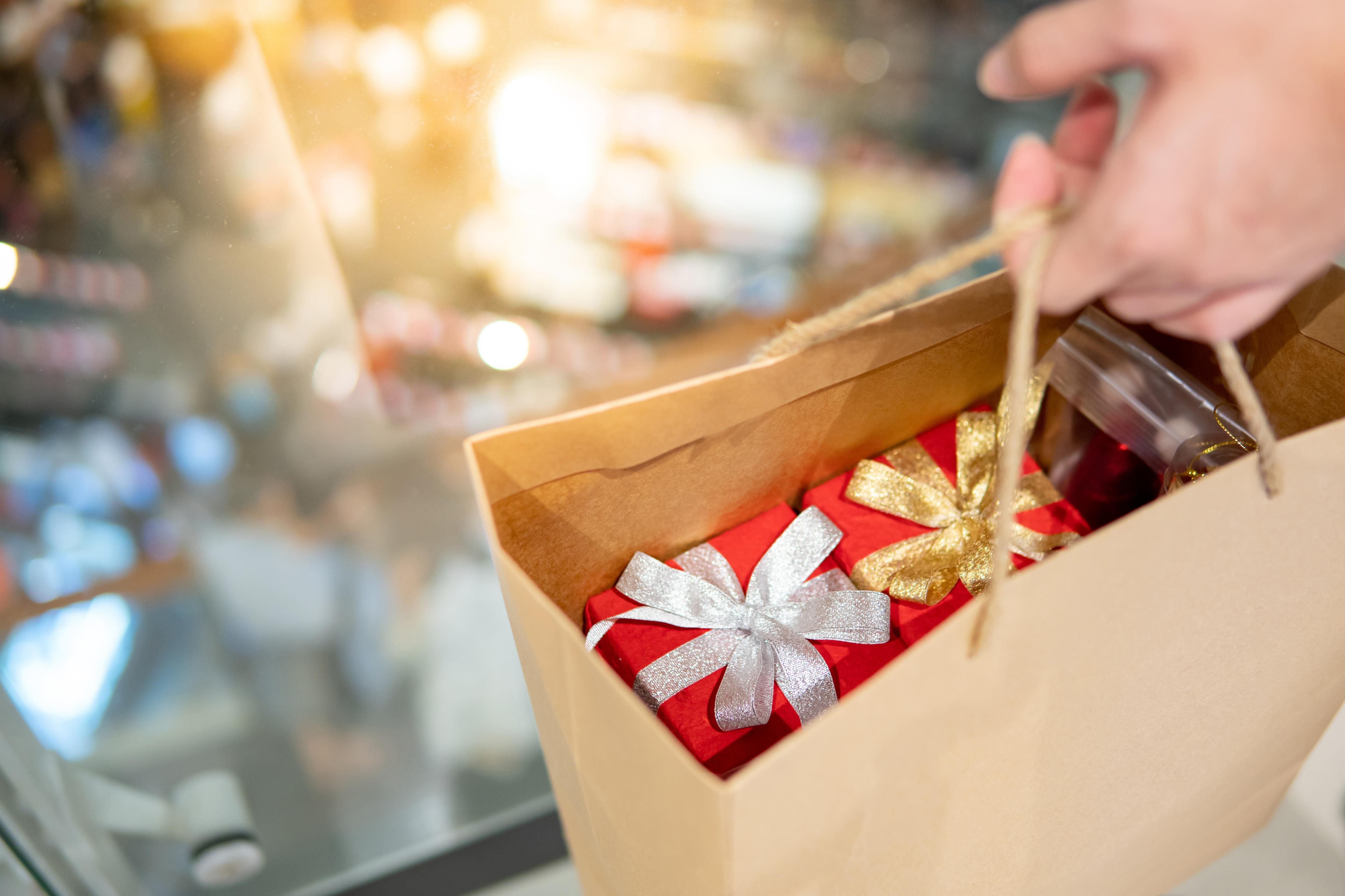 A hand holding a brown paper shopping bag filled with red gift boxes wrapped with shiny silver and gold bows.