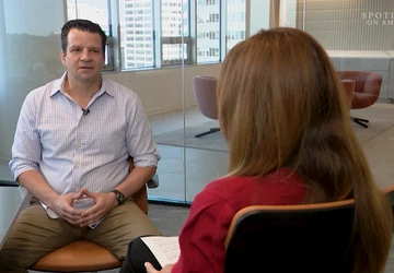 John Gray is seated in a modern glass-walled office, engaged in a conversation with a notepad visible in the foreground.