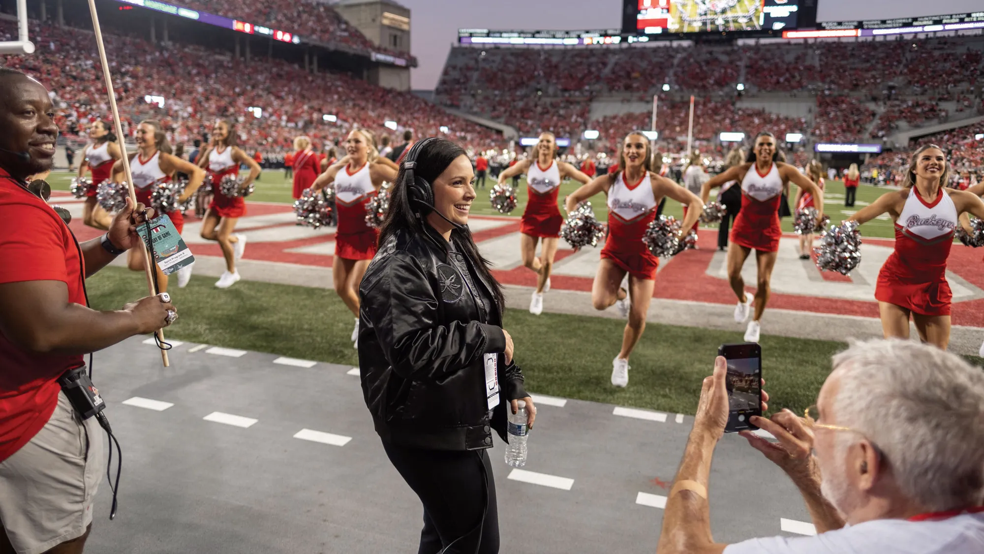 Melissa McGhee stands in front of Ohio State cheerleaders performing on a football field with a large crowd in the stadium; people on the sideline capture the moment.