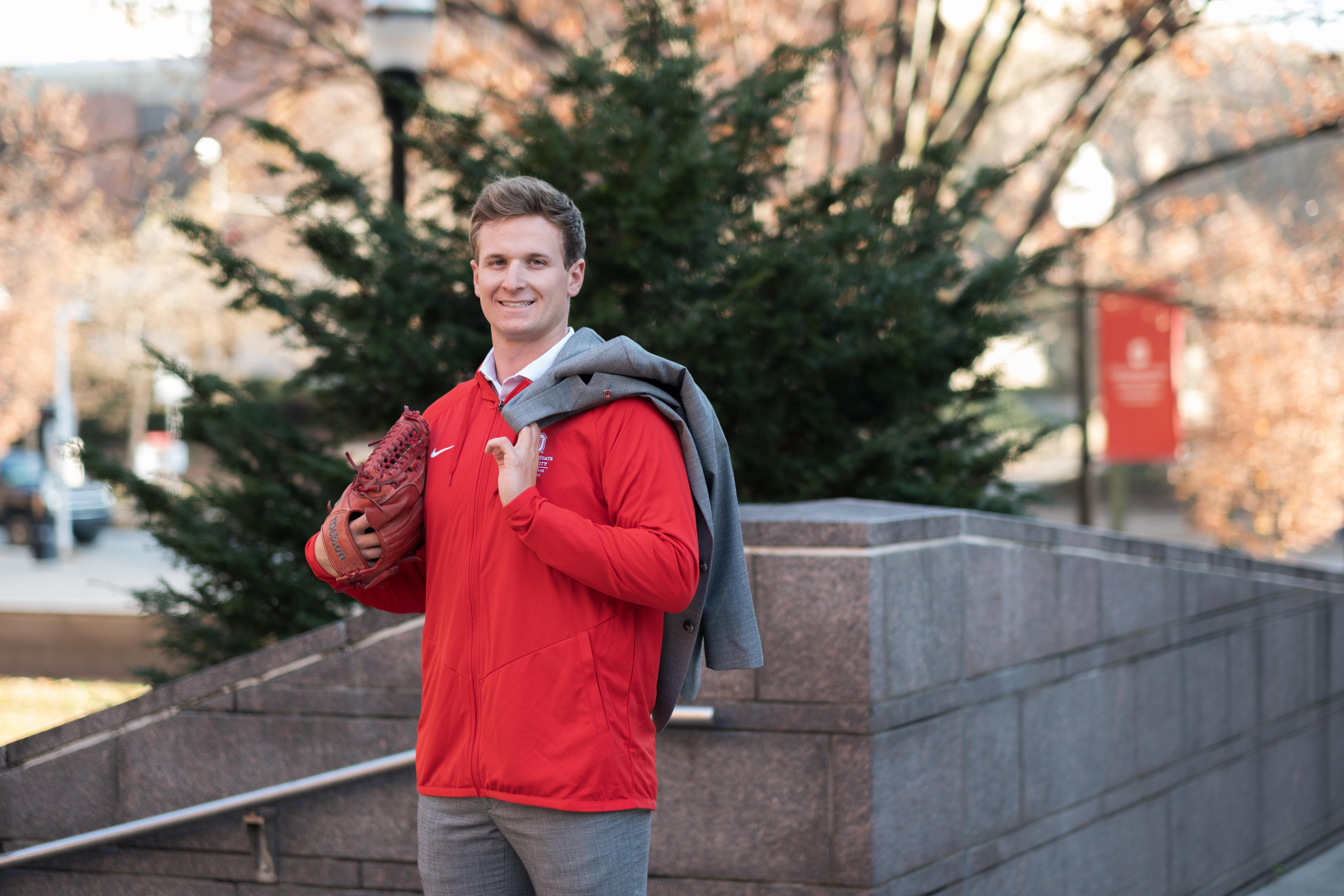 Luke Carell standing outside with red pullover, gray suit pants, red baseball glove on hand and gray suit coat draped over shoulder