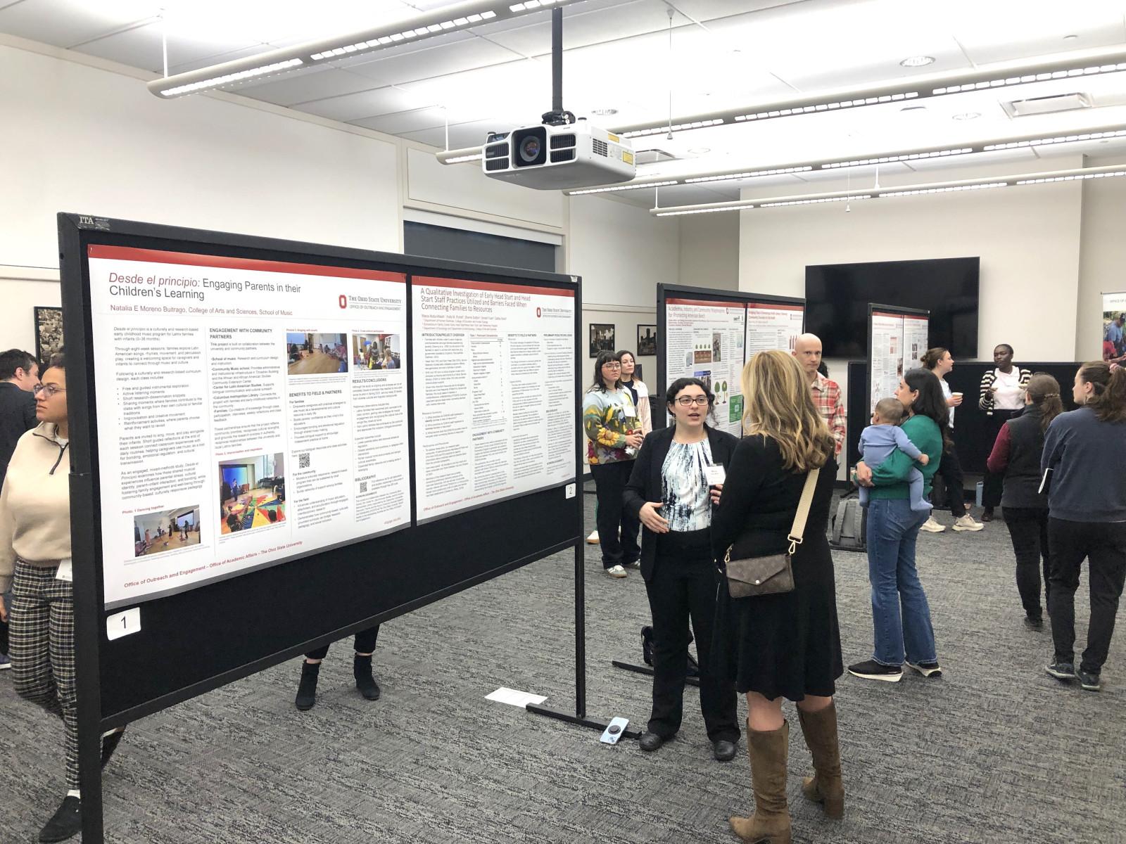 People in a conference room viewing and discussing large research posters displayed on black stands, with multiple posters arranged in rows and a ceiling-mounted projector visible above.