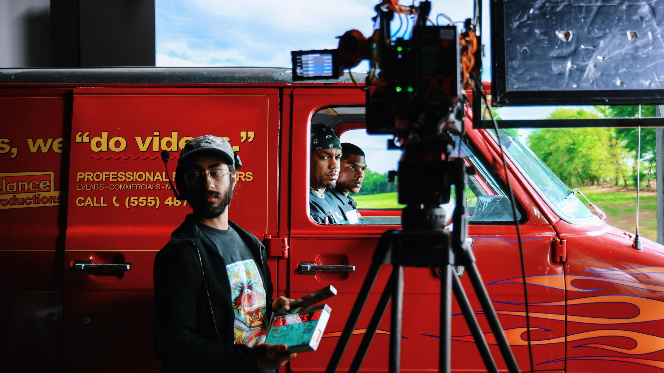 A cameraman stands beside a movie camera in front of a van with two actors inside on the set of a TV pilot.