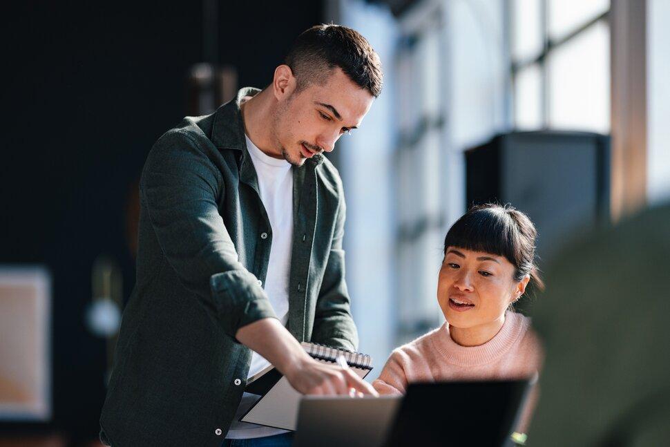 Two colleagues collaborating at a laptop, with one person pointing at the screen while discussing notes in a modern office setting.