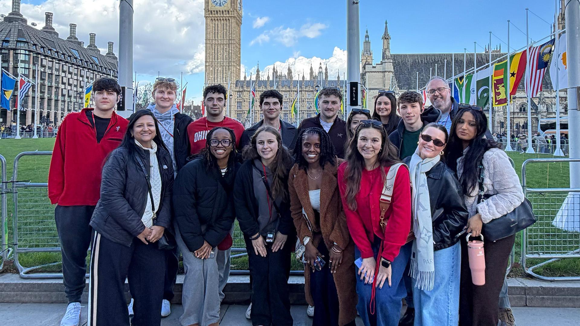 A group of Ohio State students posing together near Big Ben and the Houses of Parliament in London, with international flags and a sunny sky in the background.