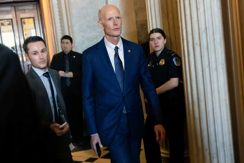 Rick Scott is escorted through the halls of a Capitol building.