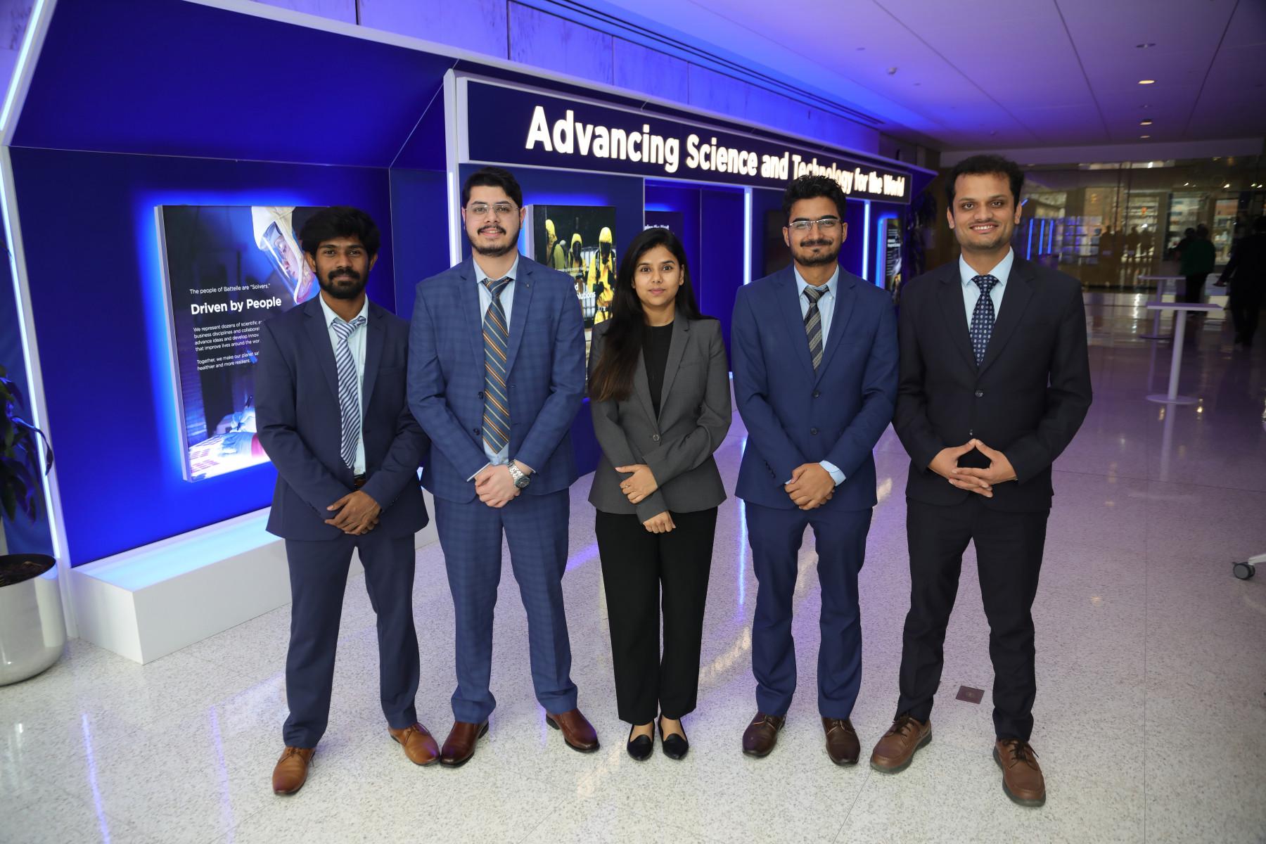 A team of MBA students in professional attire pose for a group photo in front of a blue, illuminated sign.