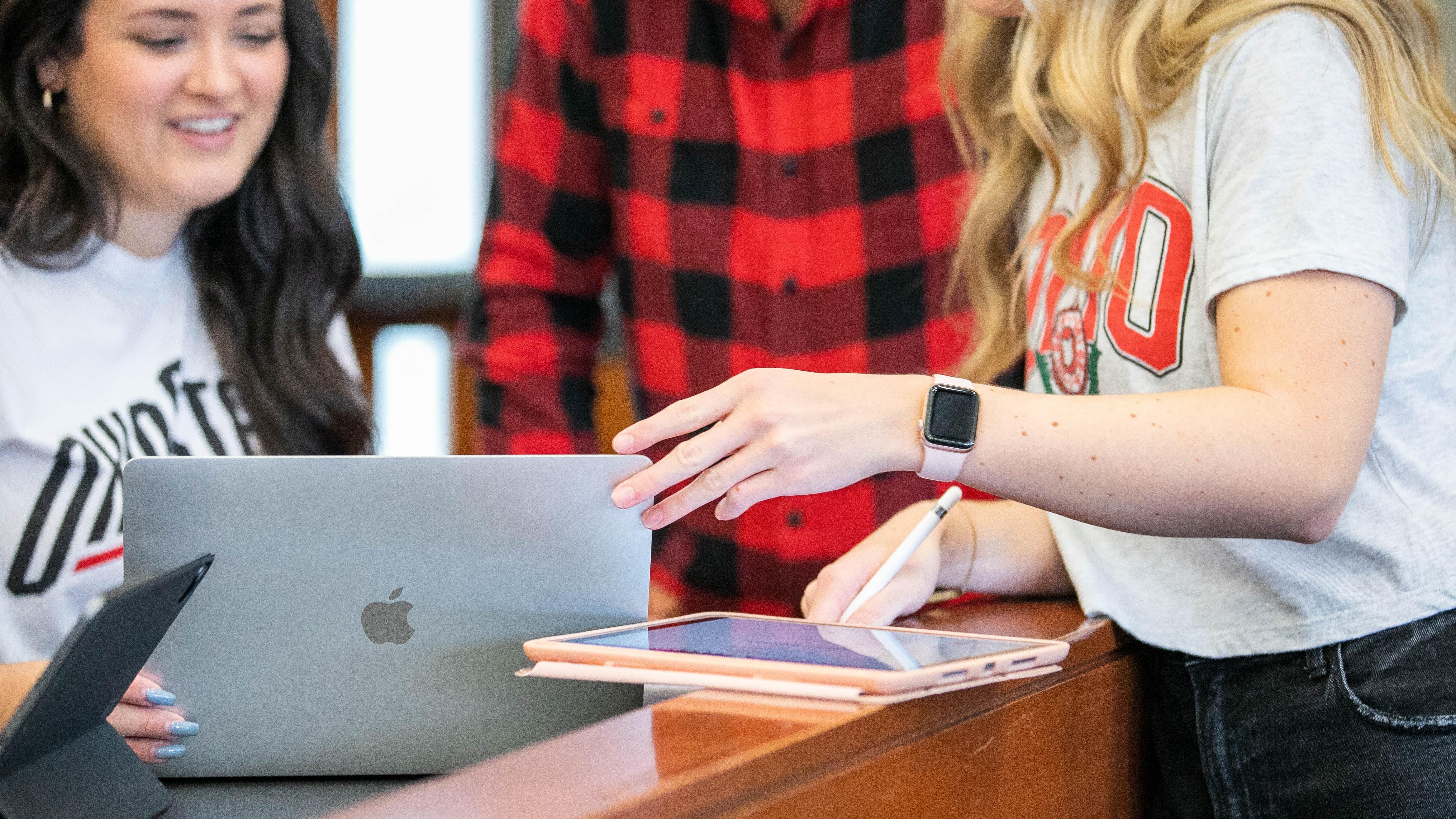 Students collaborating with a laptop and tablet, one pointing while another takes notes with a stylus.