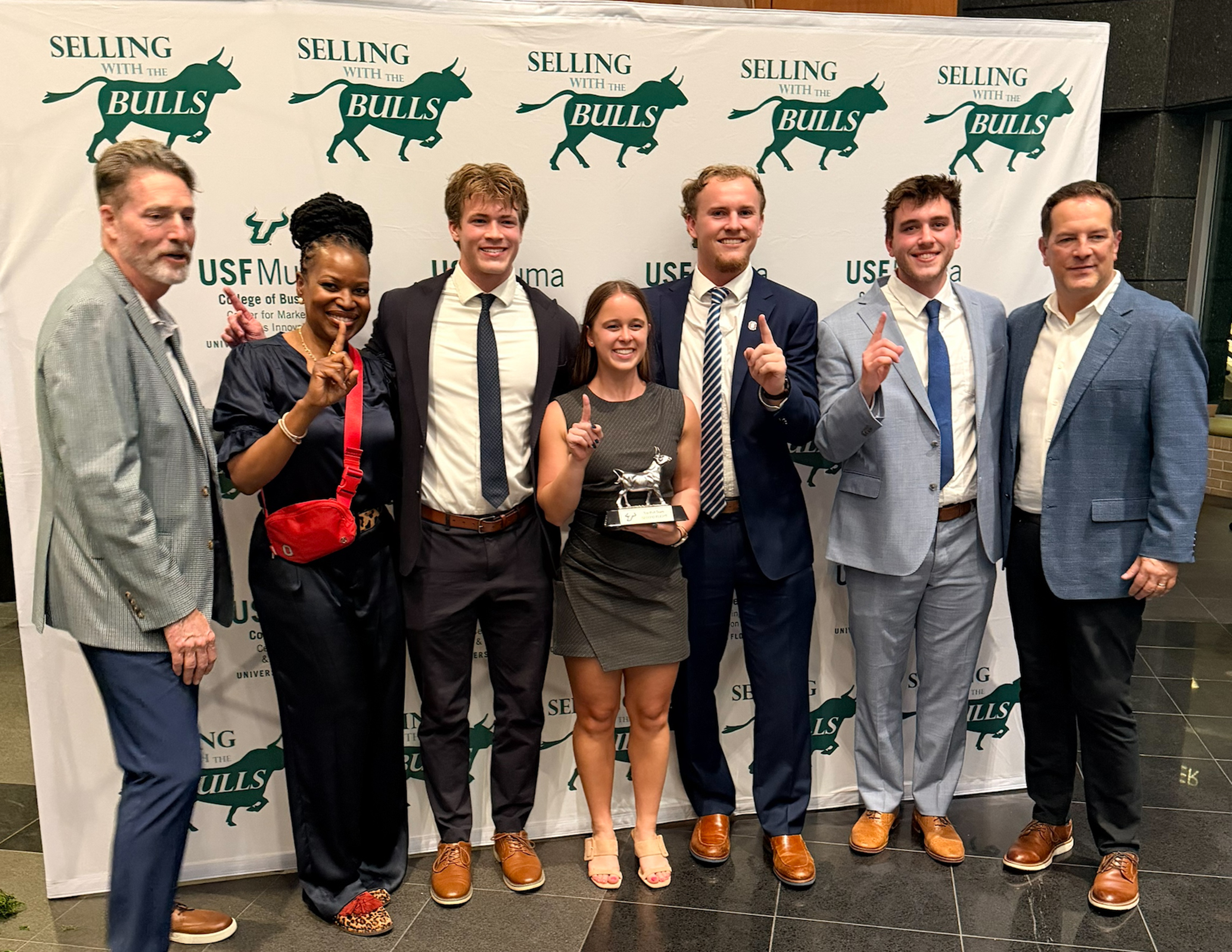 Seven people holding up finger for number one in front of a sign saying "Selling with the Bulls"