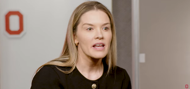 Kathy Wantuch answers questions in a black shirt in front of a Block O.