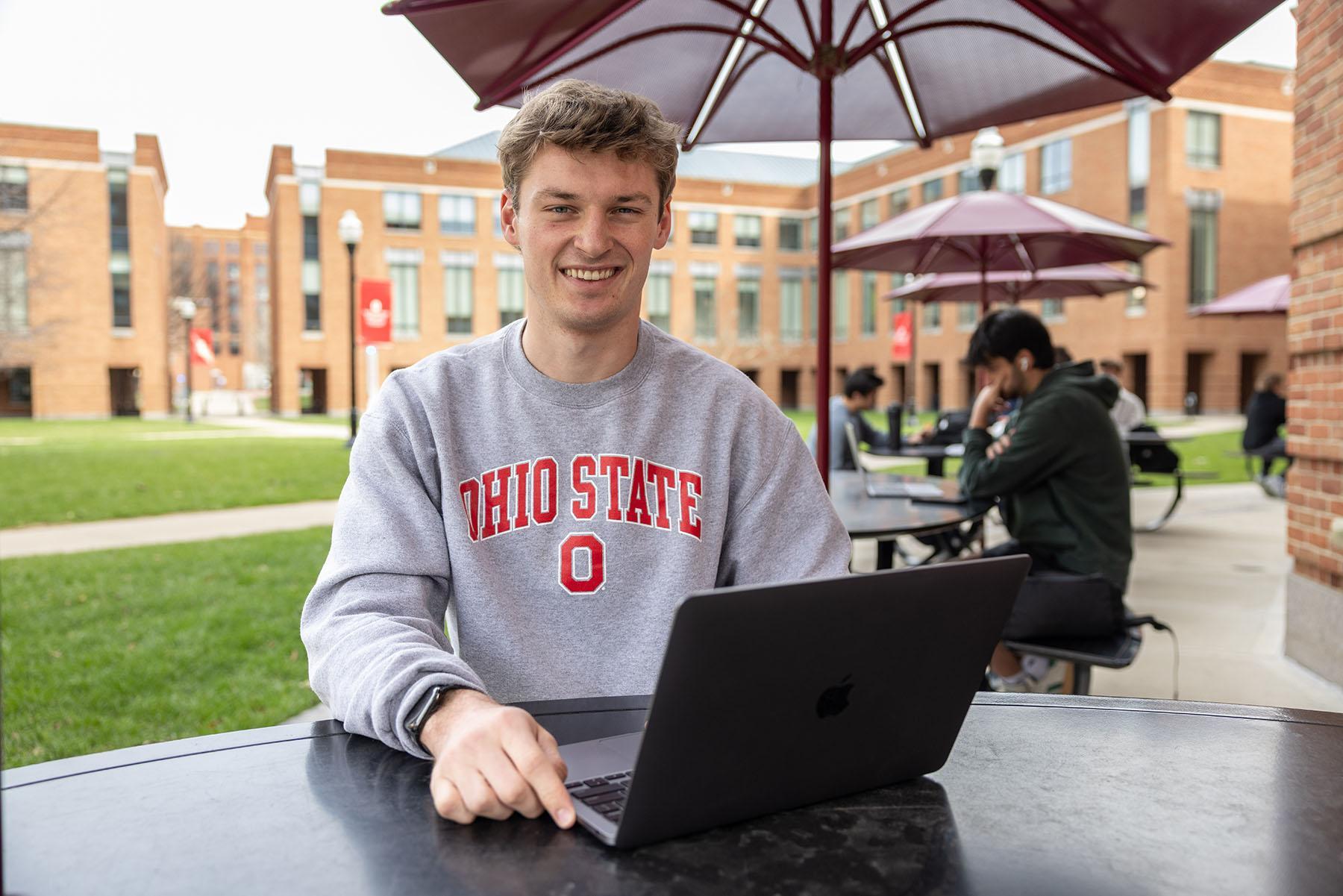 A student in a gray Ohio State sweatshirt sits outside under an umbrella in front of an open laptop.