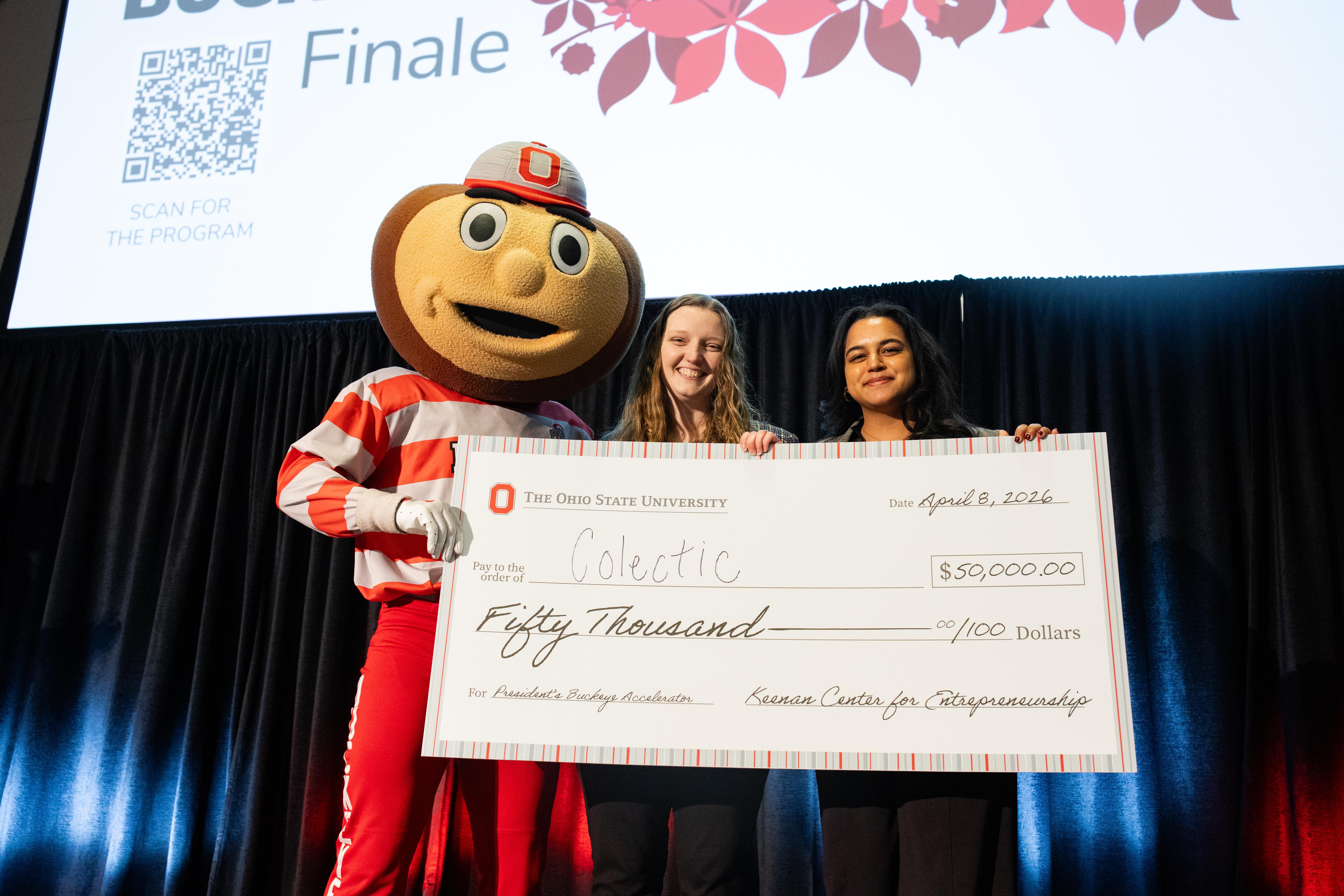 Two students pose with Brutus Buckeye and large check on stage at a pitch competition.
