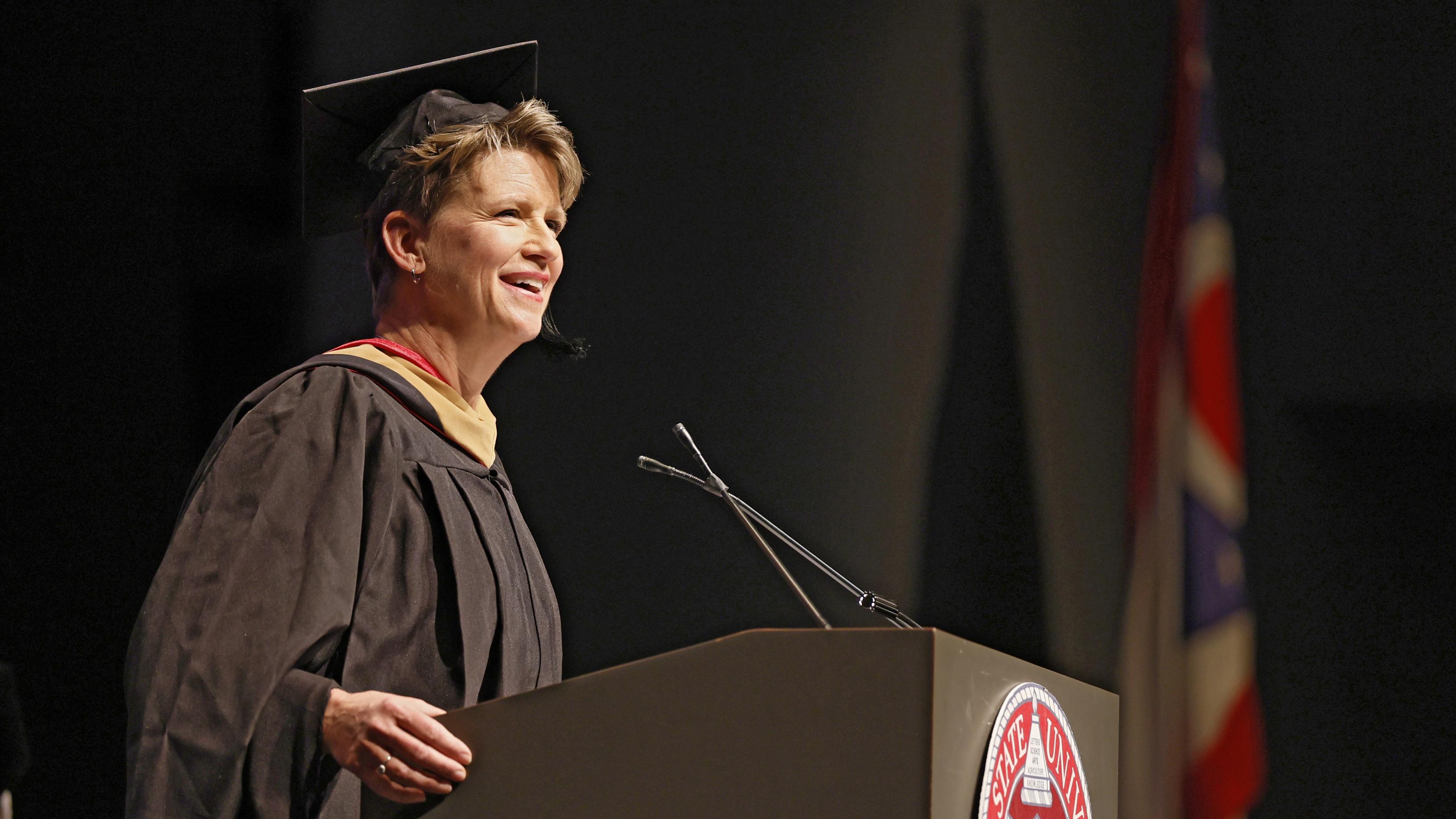 Melinda Whittington in a black cap and gown and speaking into a microphone at the podium during the Pre-Commencement Ceremony.