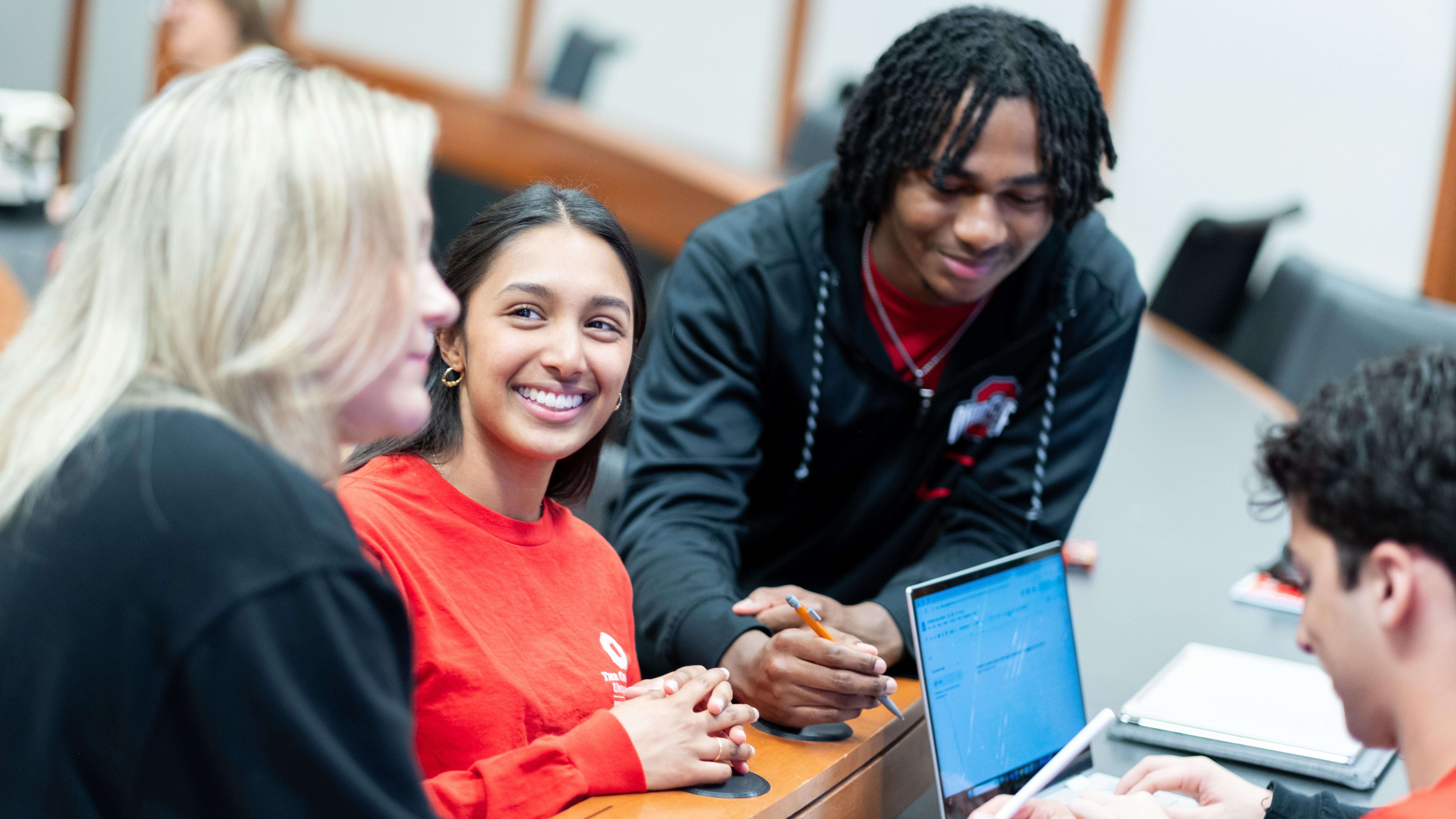 Students collaborate at a table, reviewing notes on a laptop while discussing a project in a classroom setting.