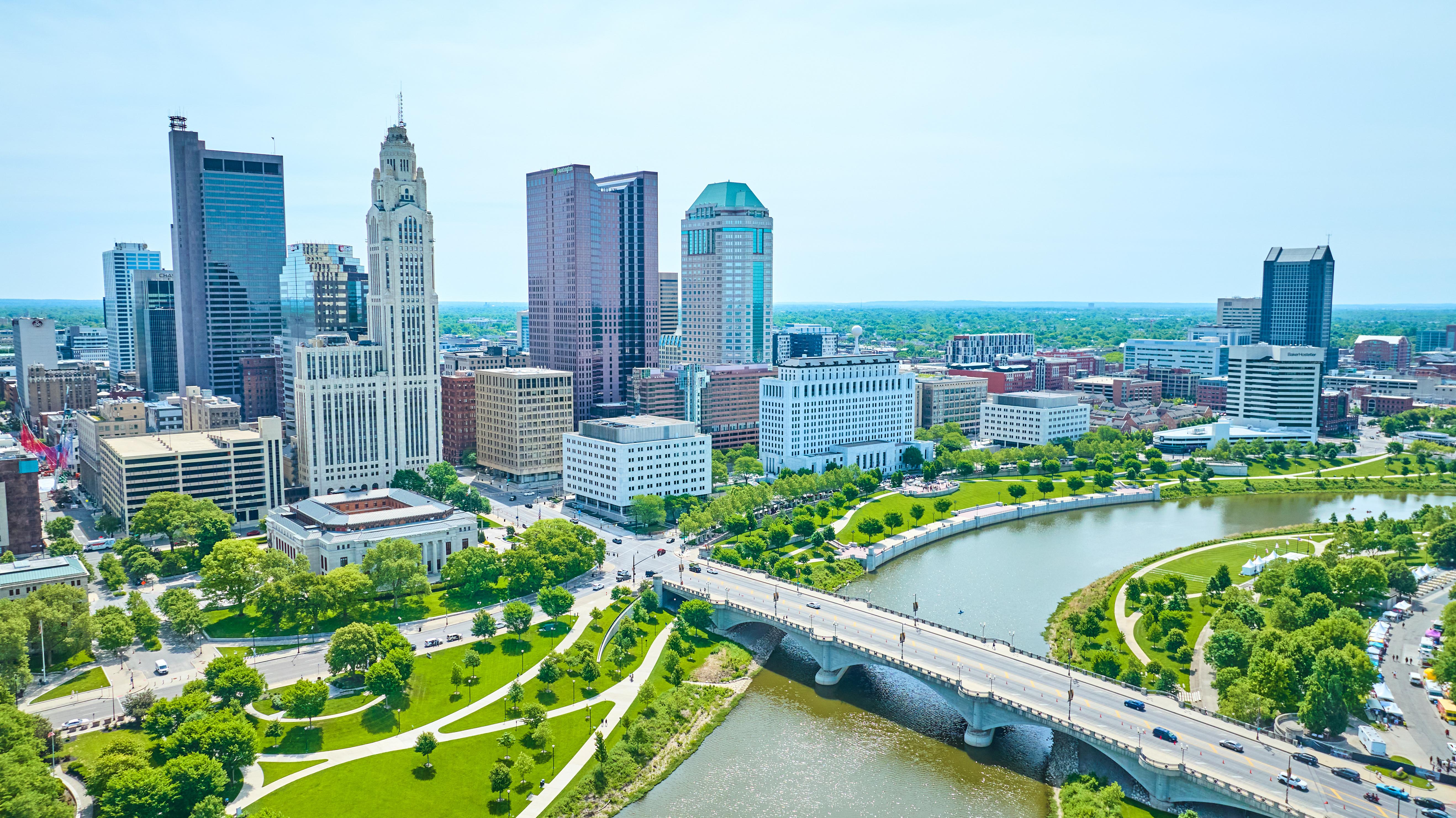 Aerial view of downtown Columbus, Ohio, featuring the city skyline, Scioto River, and surrounding green parkland on a sunny day.