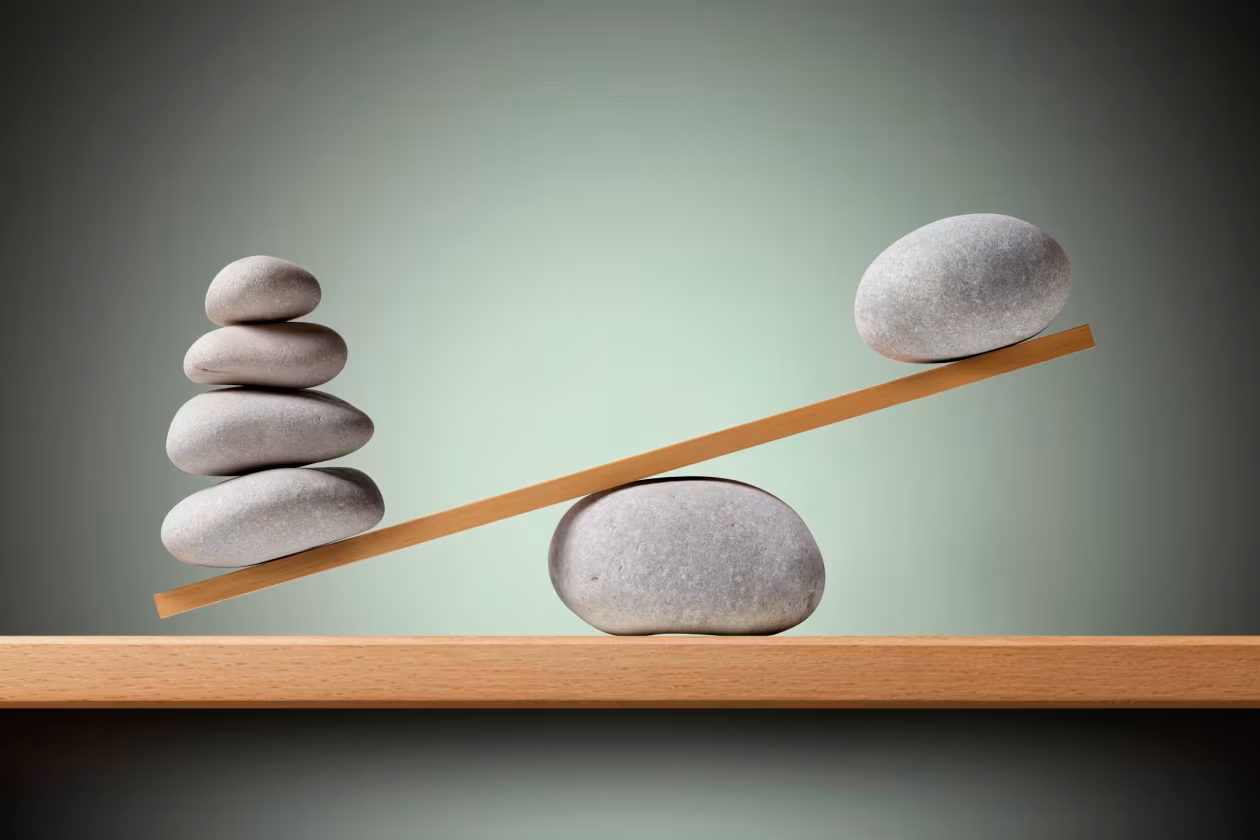 Wooden balance beam tipped under a stack of four stones on one side and a single stone on the other, illustrating imbalance or competing weight.