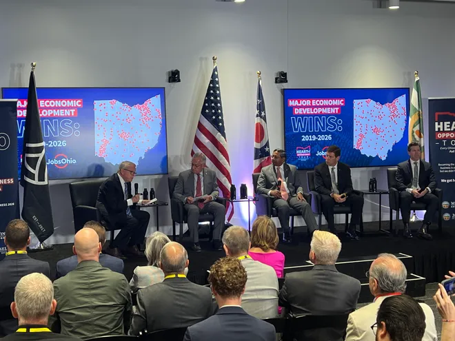 Audience watches a panel discussion on economic development in Ohio, with state flags and presentation screens in the background.