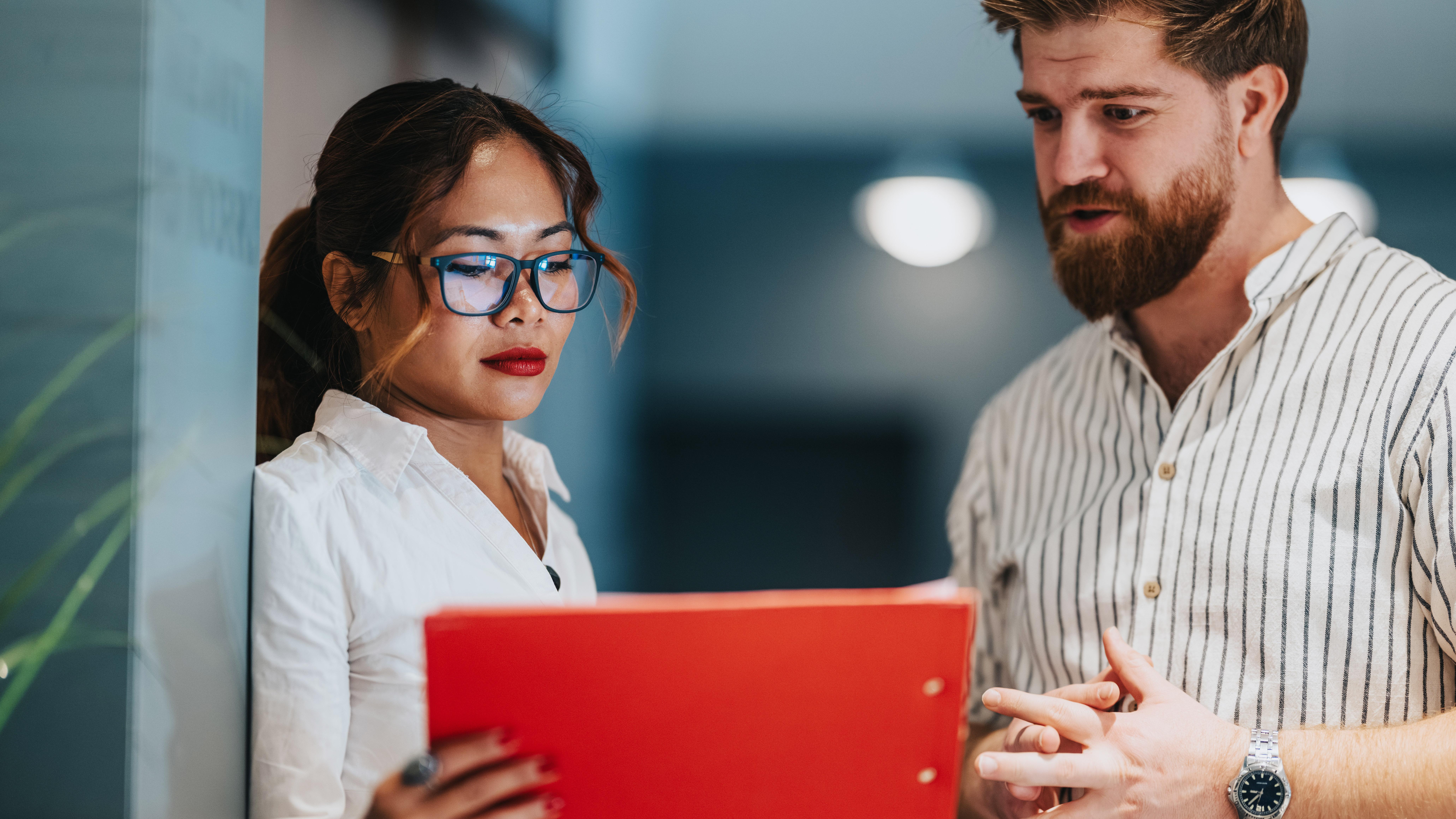 Two professionals review a red folder while discussing documents in an office setting.
