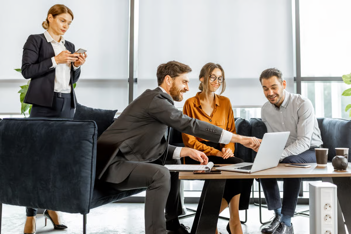 Business professionals gathered around a laptop in a modern office meeting area, reviewing information together.