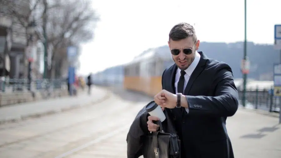 Person in a business suit at a train platform checking a wristwatch while holding a briefcase and travel mug.