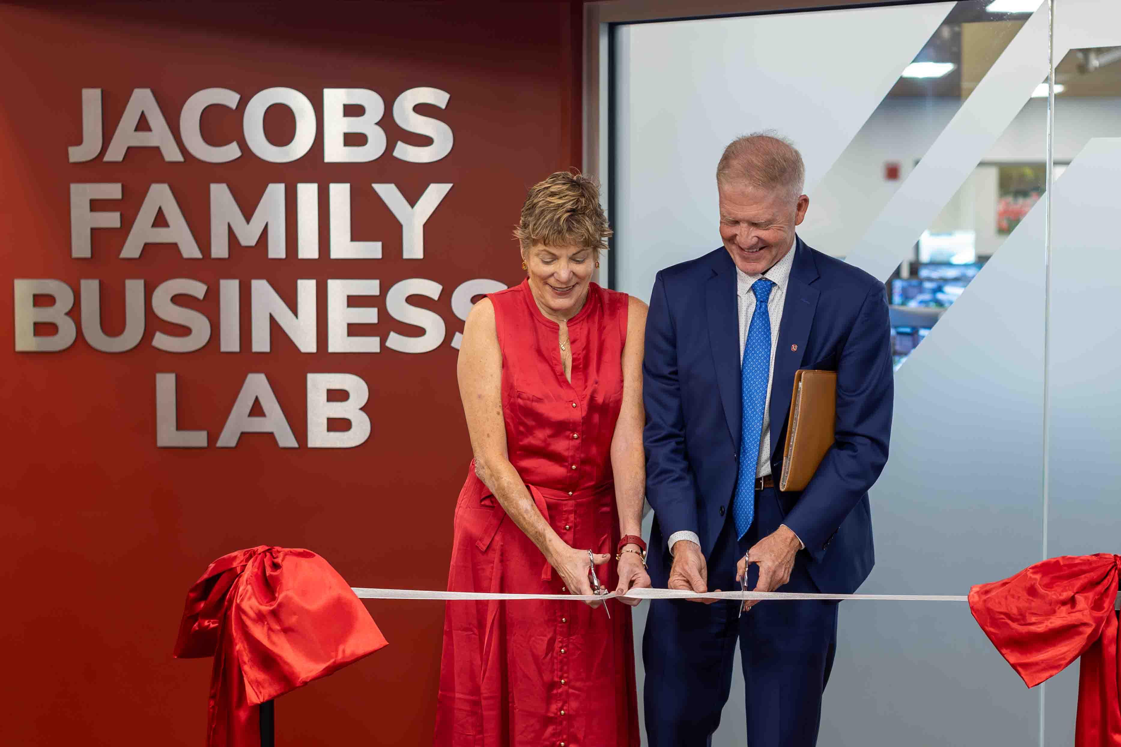 Brenda Jacobs in a red dress and Dave Jacobs in a blue suit each hold a pair of scissors and cut into a ribbon in front of the Jacobs Family Business Lab.