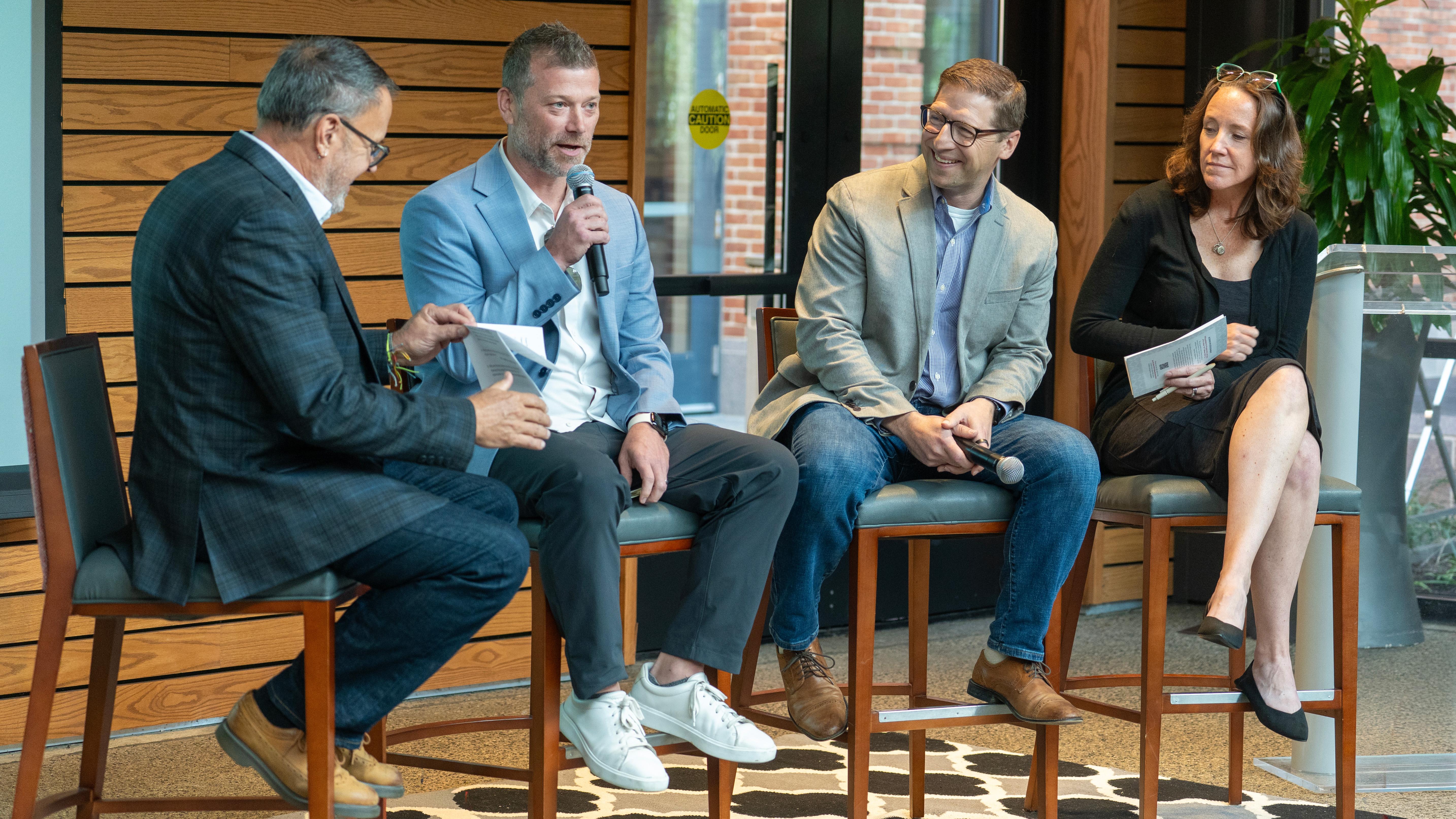 Three men and one woman sit on high stools as part of a panel discussion, with one man holding a microphone.