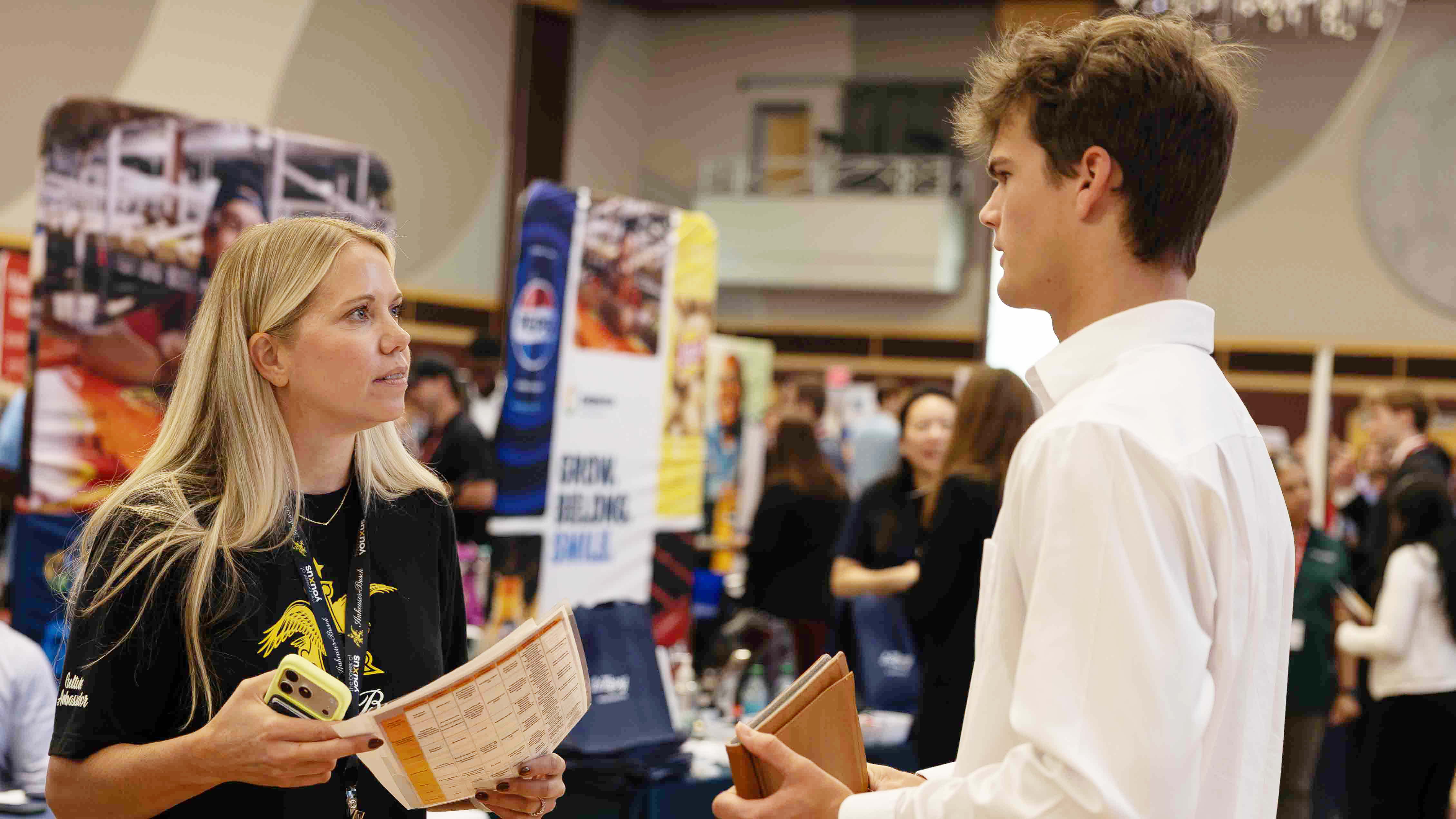 Female job recruiter in black company tee shirt greets a male student in a white dress shirt and holding a folder.