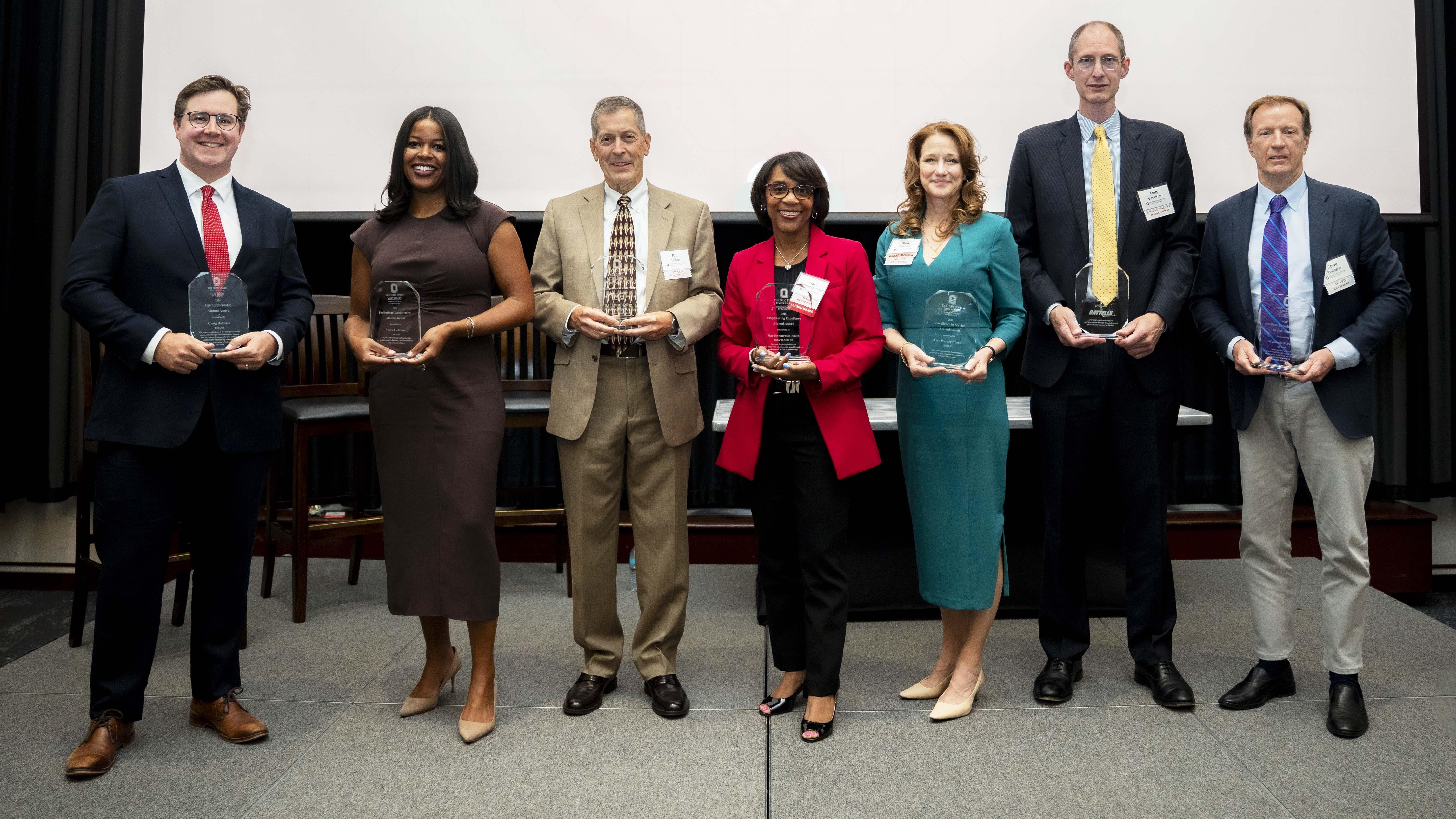 Seven men and women in business dress stand in a line holding glass awards.