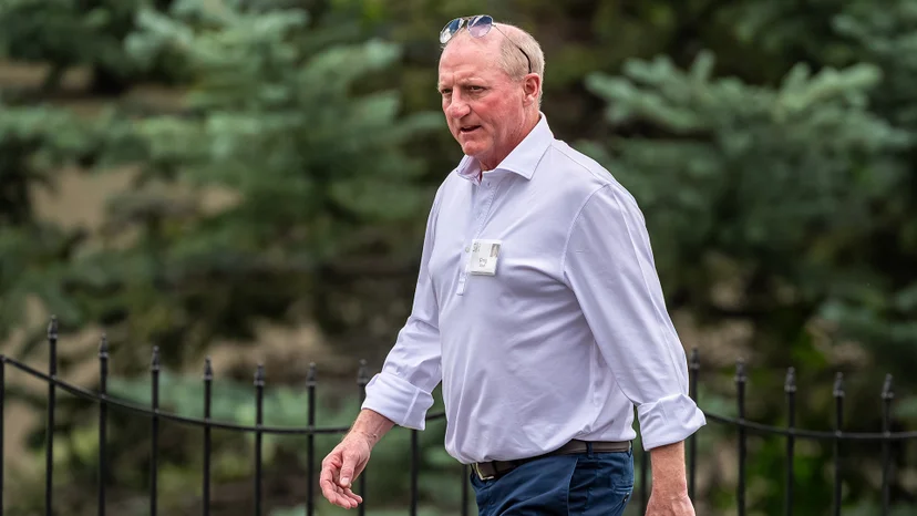 Berkshire Hathaway CEO Greg Abel, wearing a light shirt, walks outdoors beside a black metal fence and evergreen trees.