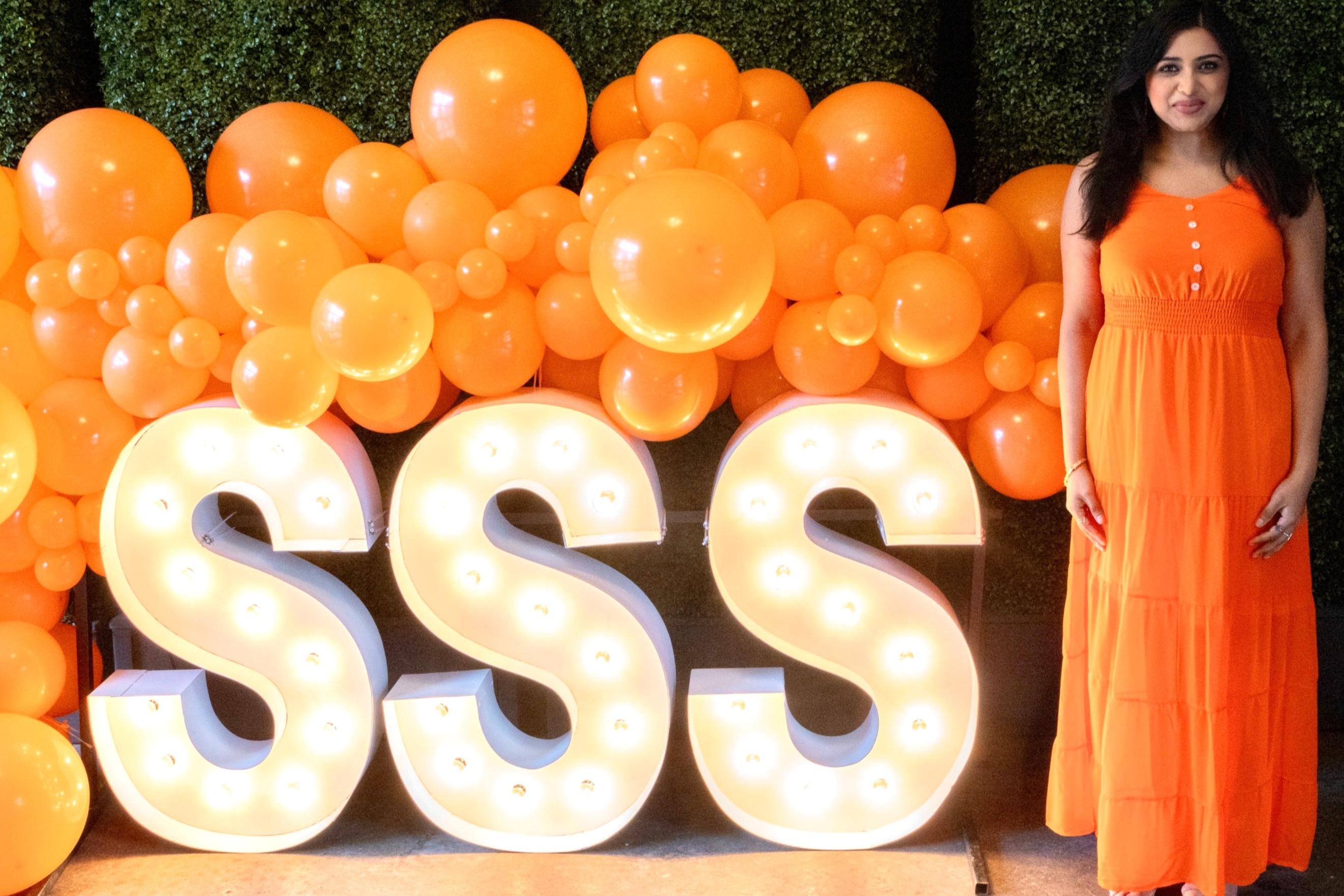 Neethi Johnson in an orange dress standing to the right of a lighted SSS sign and decorative orange balloons.